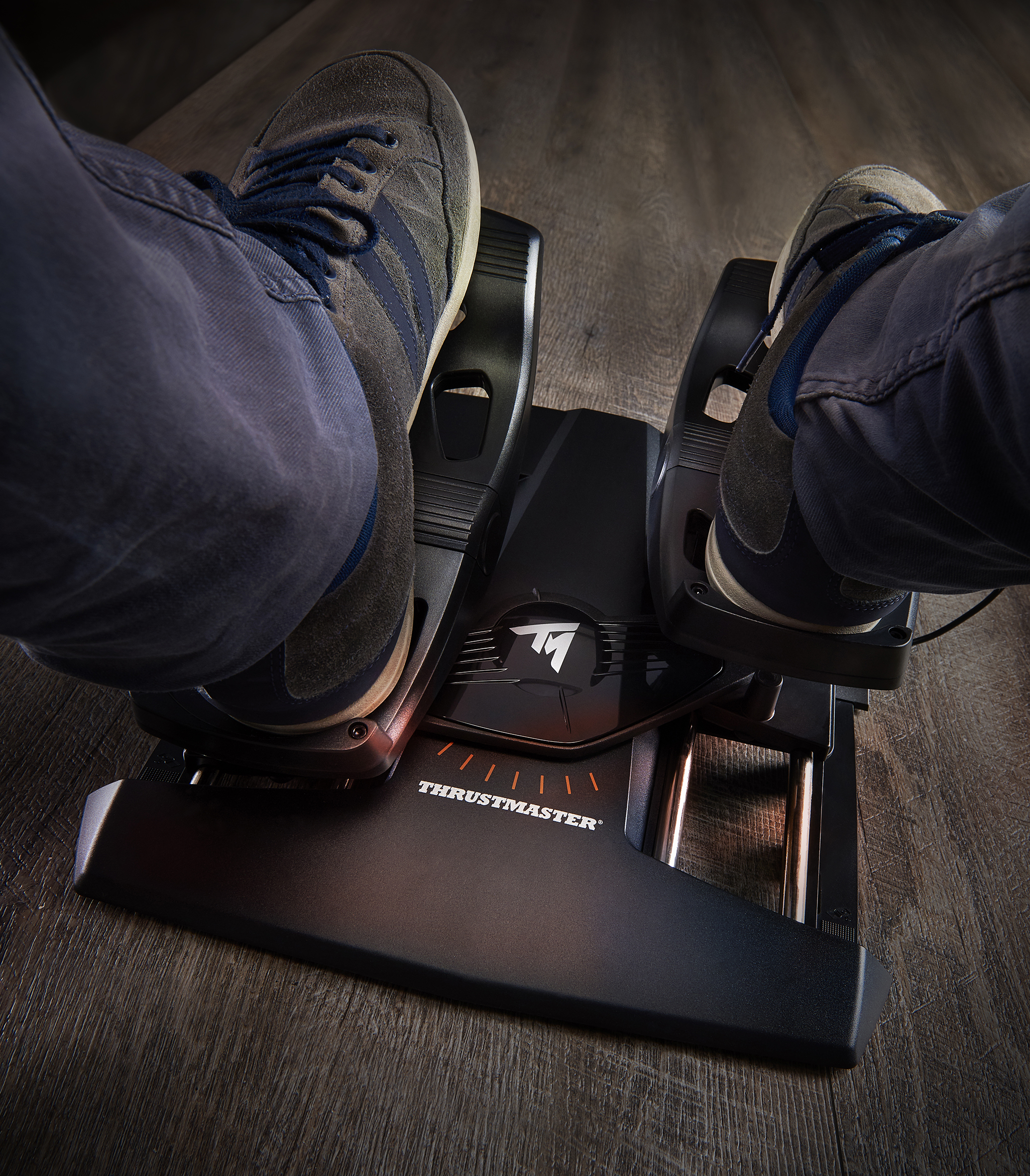 Feet on a Thrustmaster pedal set. Gray shoes, blue jeans, and wood floor. Black pedals and the Thrustmaster logo are visible.