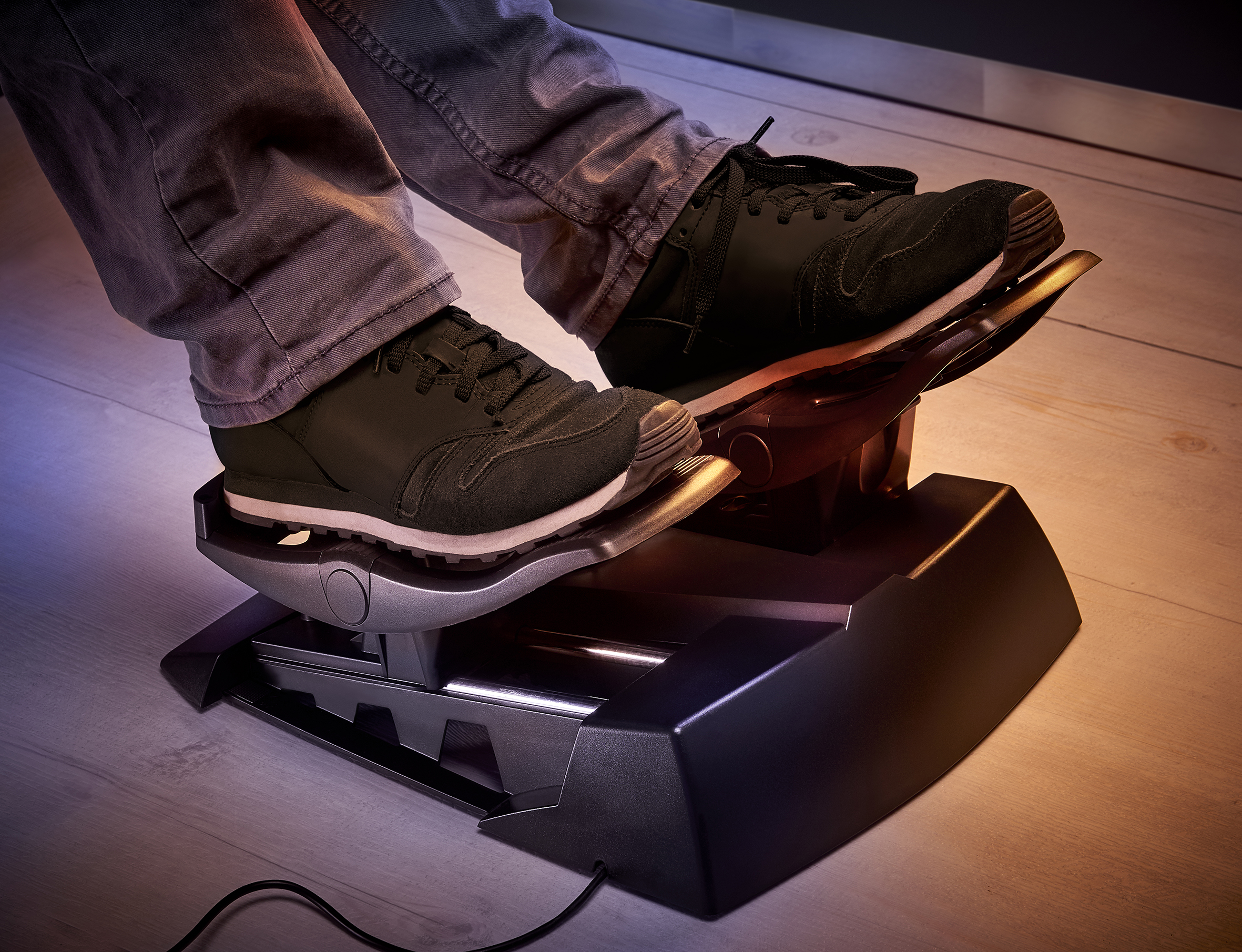 Person's feet on a black foot pedal controller. The person is wearing jeans and black sneakers. The floor is wooden.