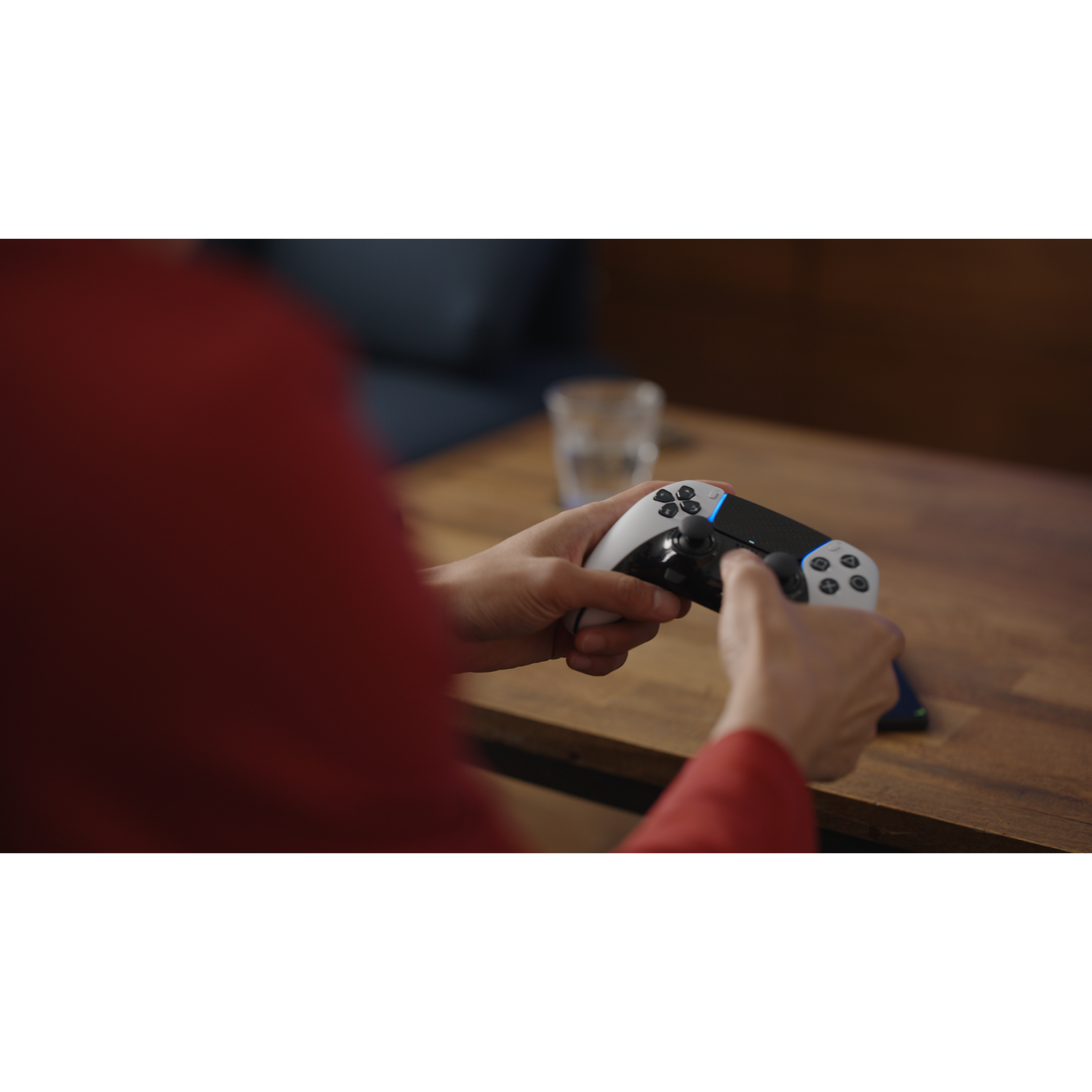 Handen die een zwart-witte gamecontroller vasthouden op een houten tafel. Een glas water op de achtergrond.