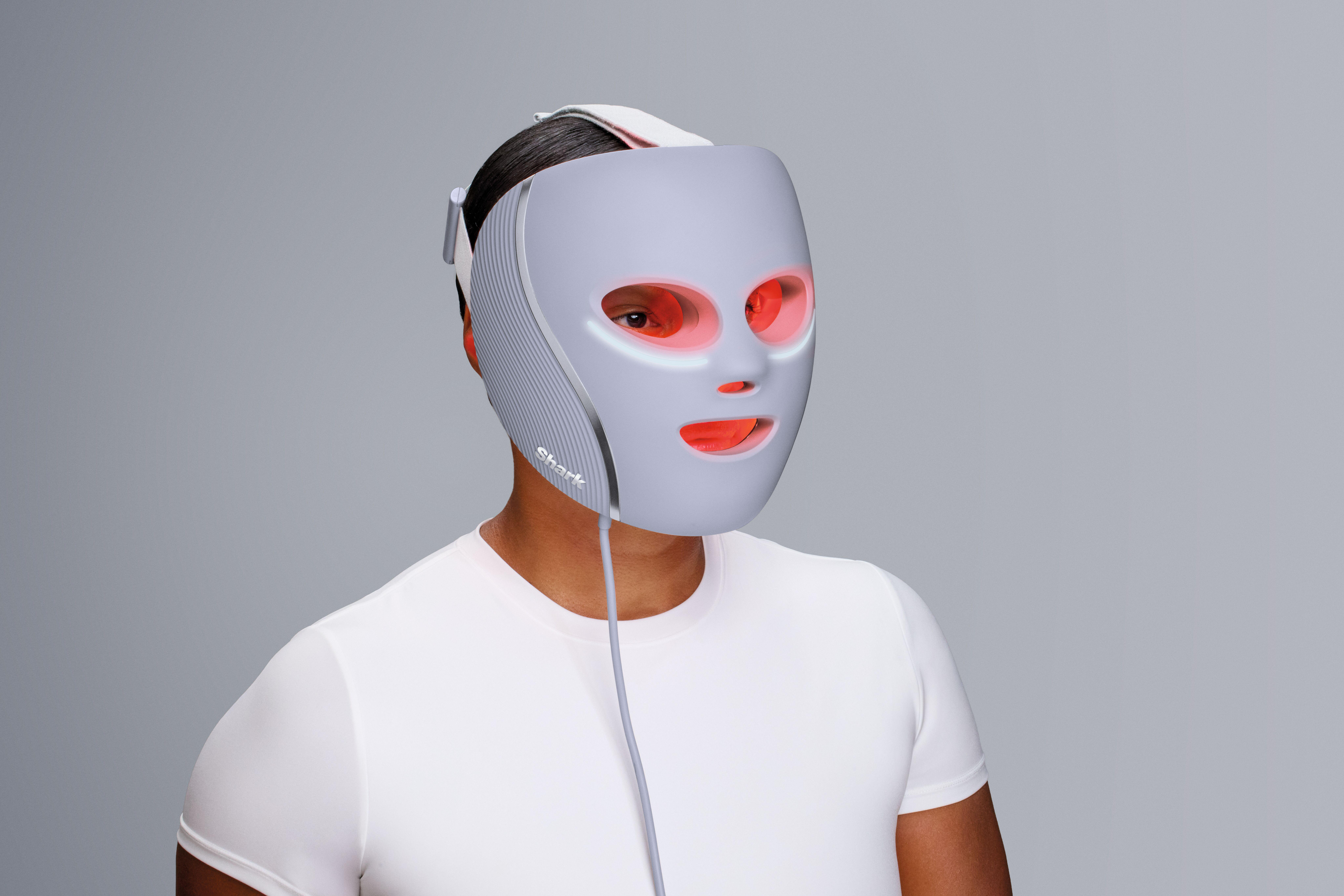 Person trägt graue LED-Gesichtsmaske. Augen und Mund sind sichtbar. Die Person trägt ein weißes Hemd vor grauem Hintergrund.