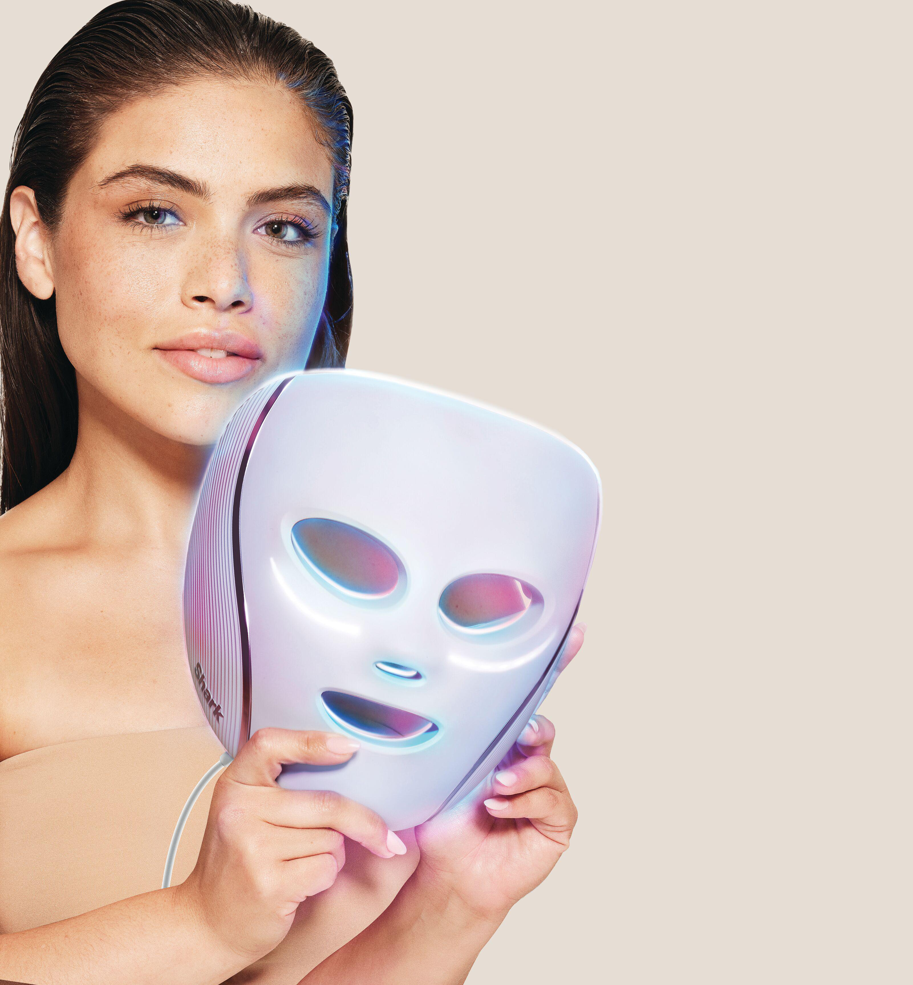 Eine Frau hält eine weiße Gesichtsmaske mit Lichtern. Sie hat dunkle Haare und schaut in die Kamera. Der Hintergrund ist beige.