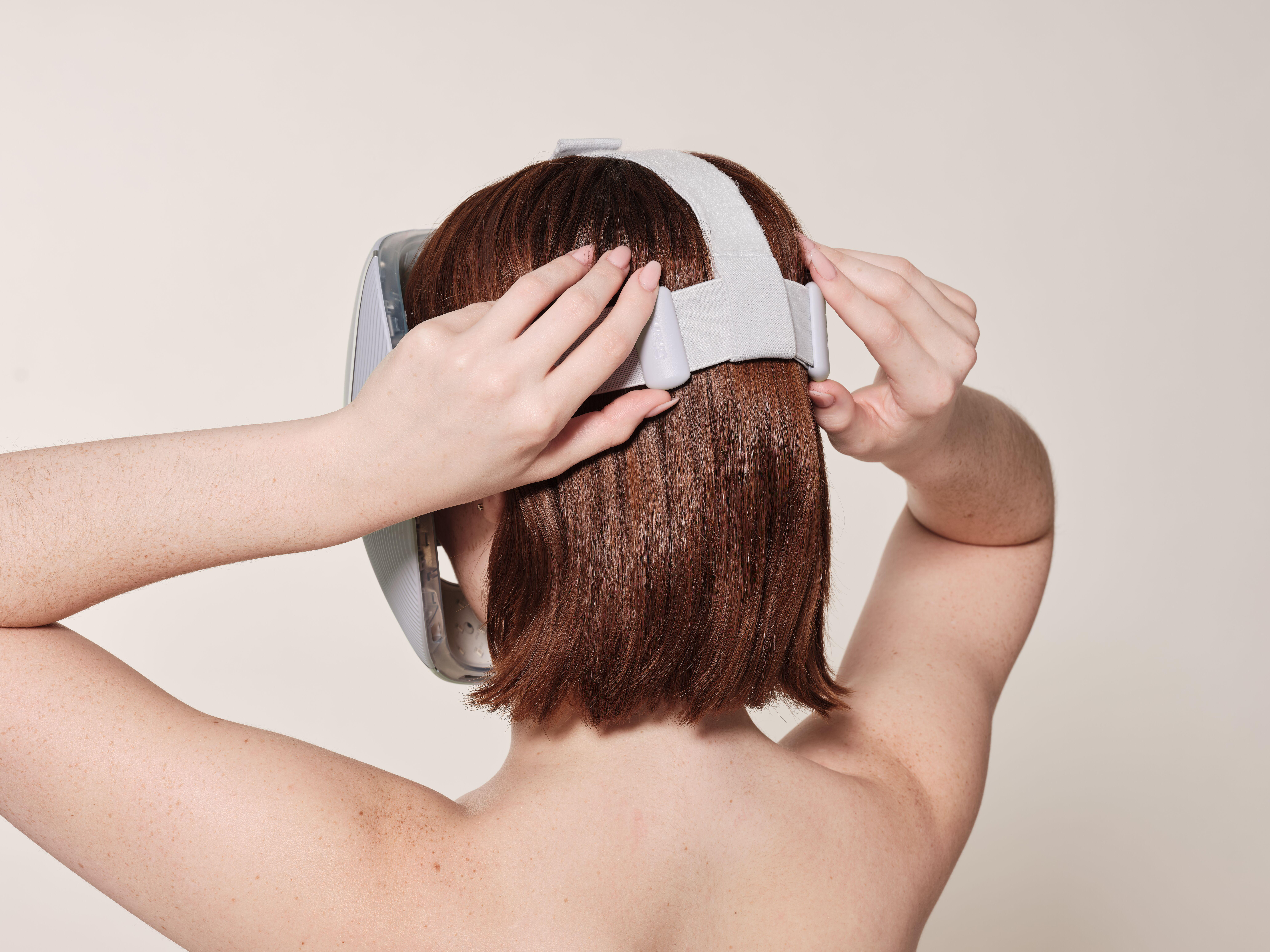 Eine Person mit kurzen, rotbraunen Haaren setzt ein VR-Headset auf. Rückansicht. Beigefarbener Hintergrund. Hände passen den Riemen an.
