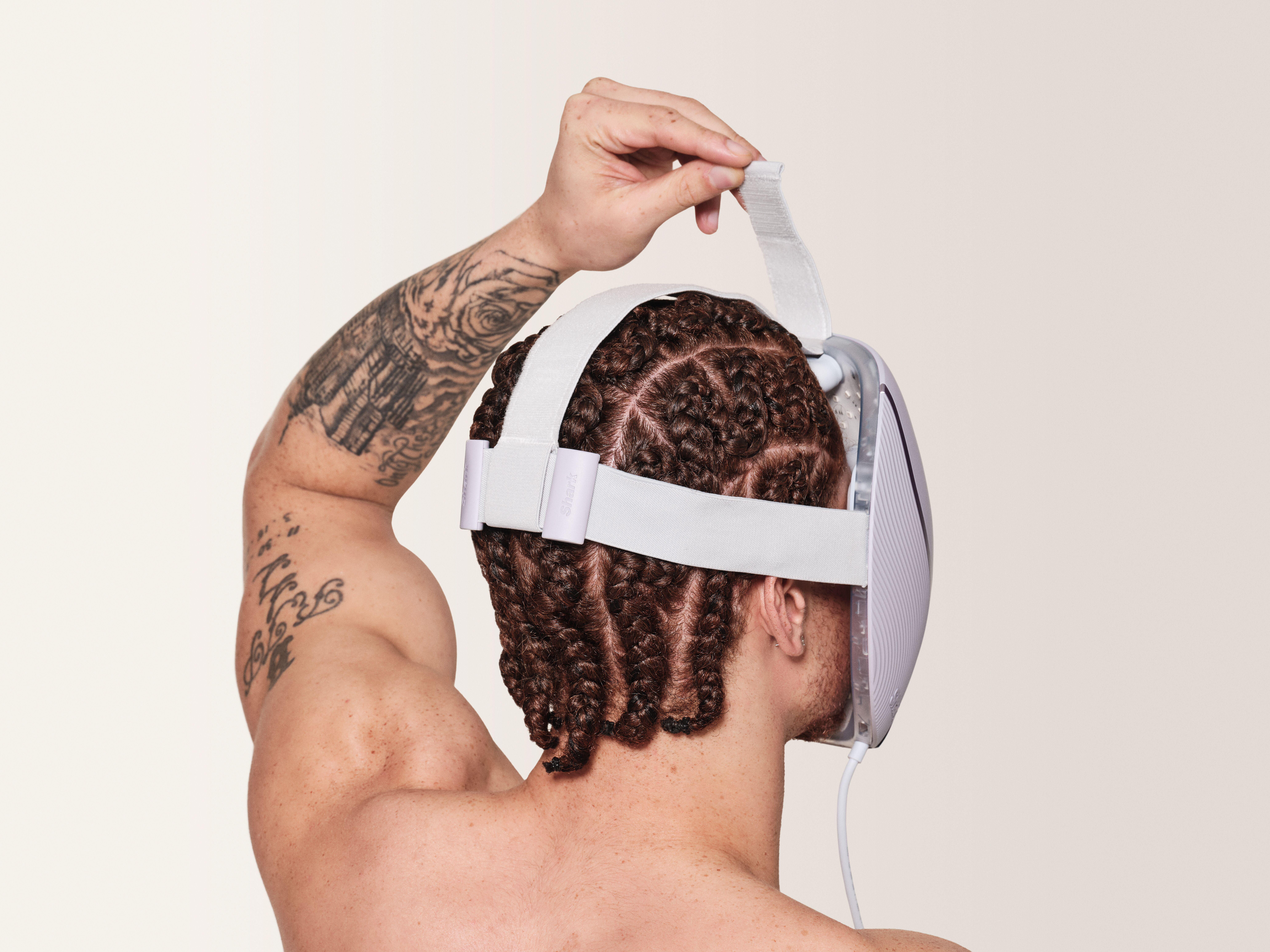 Ein Mann mit Zöpfen, der eine hellfarbige Gesichtsmaske trägt und den Riemen einstellt. Er hat Tätowierungen auf dem Arm.