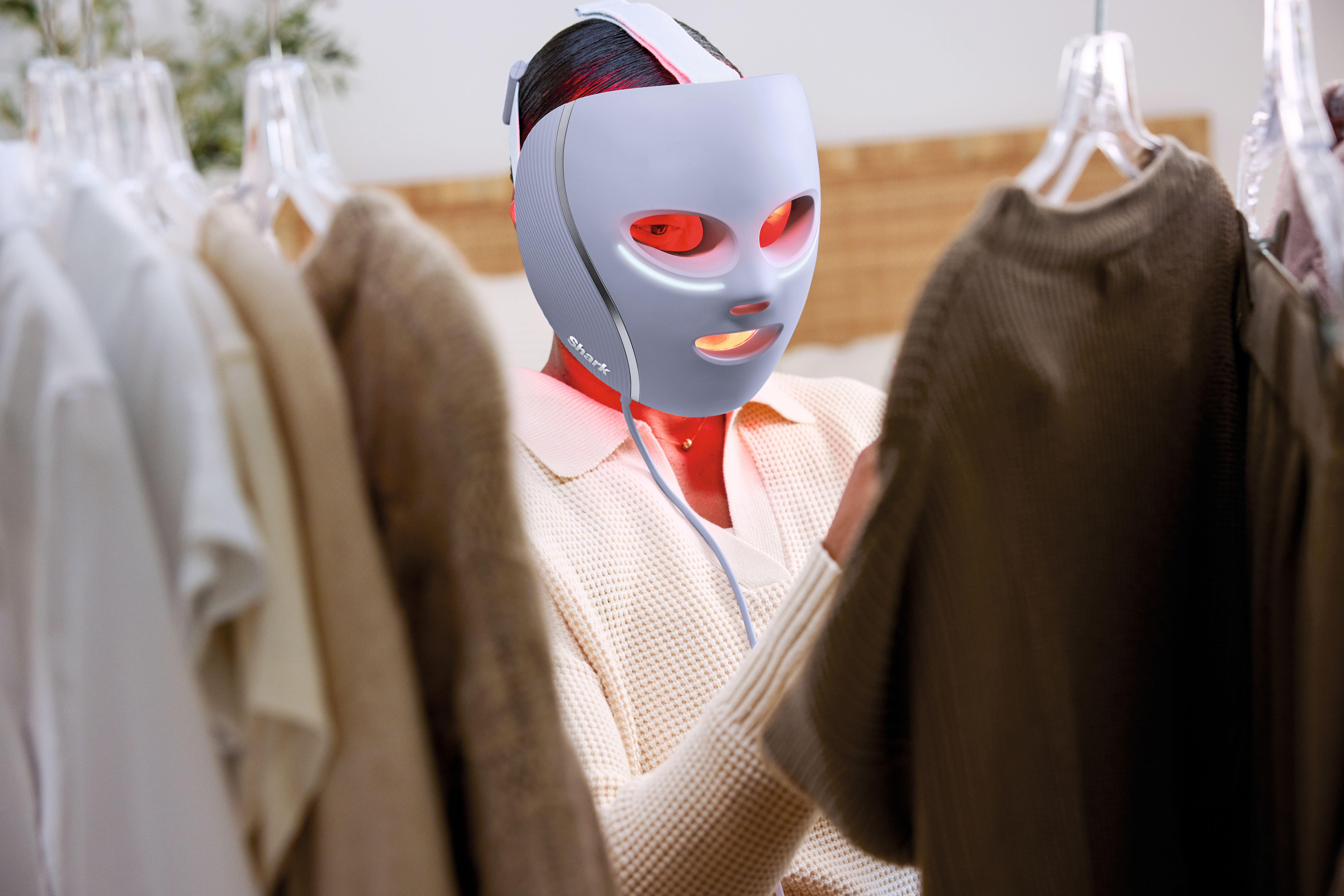 Eine Person mit einer Lichttherapiemaske steht vor Kleidung auf Kleiderbügeln. Die Maske hat rot leuchtende Lichter.