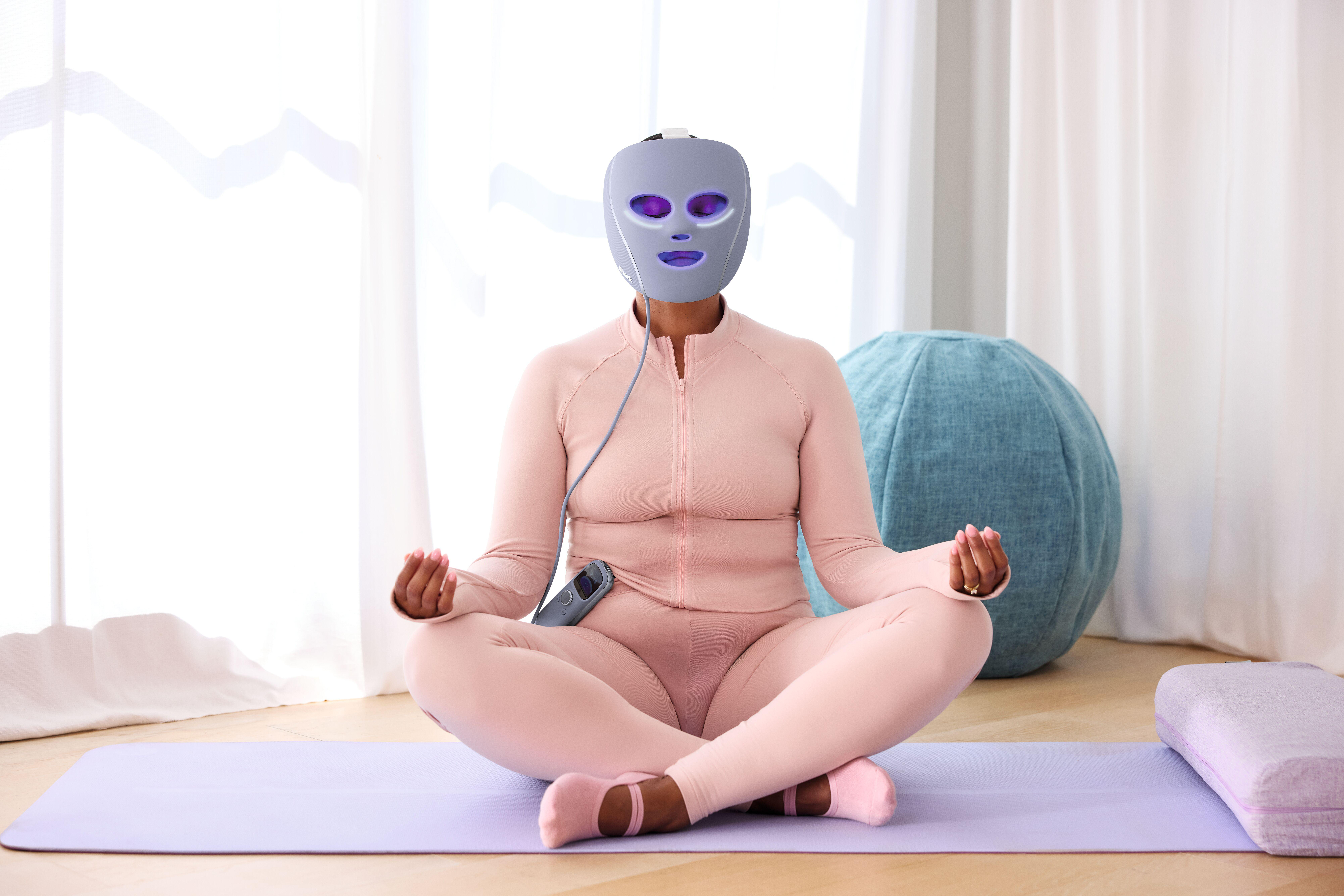 Person in rosa Anzug sitzt im Lotussitz und trägt eine Lichttherapiemaske. Blauer Sitzsack und weiße Vorhänge im Hintergrund.