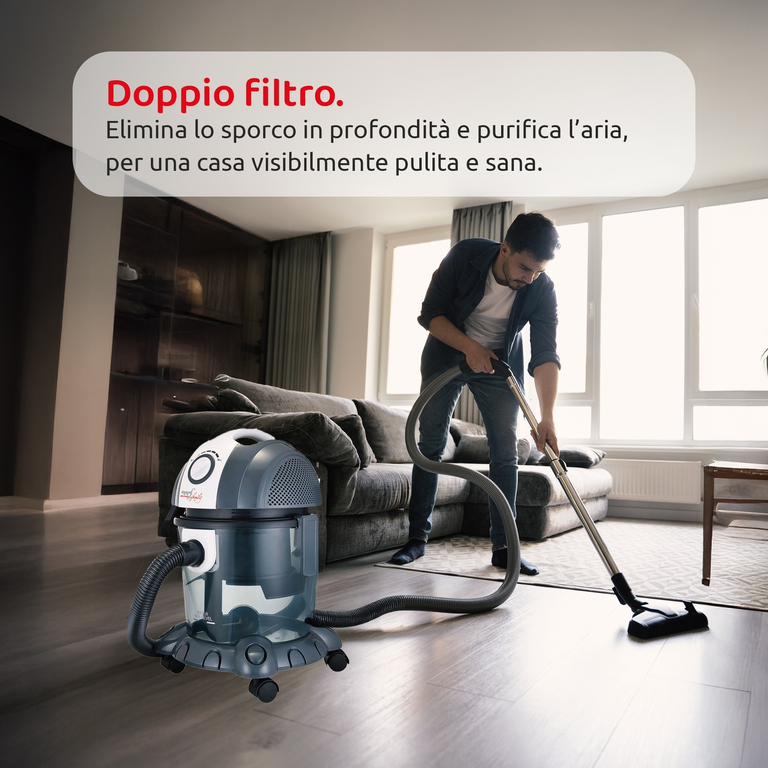 Un uomo passa l'aspirapolvere in un soggiorno. Un aspirapolvere è in primo piano e un divano e ampie finestre sullo sfondo.