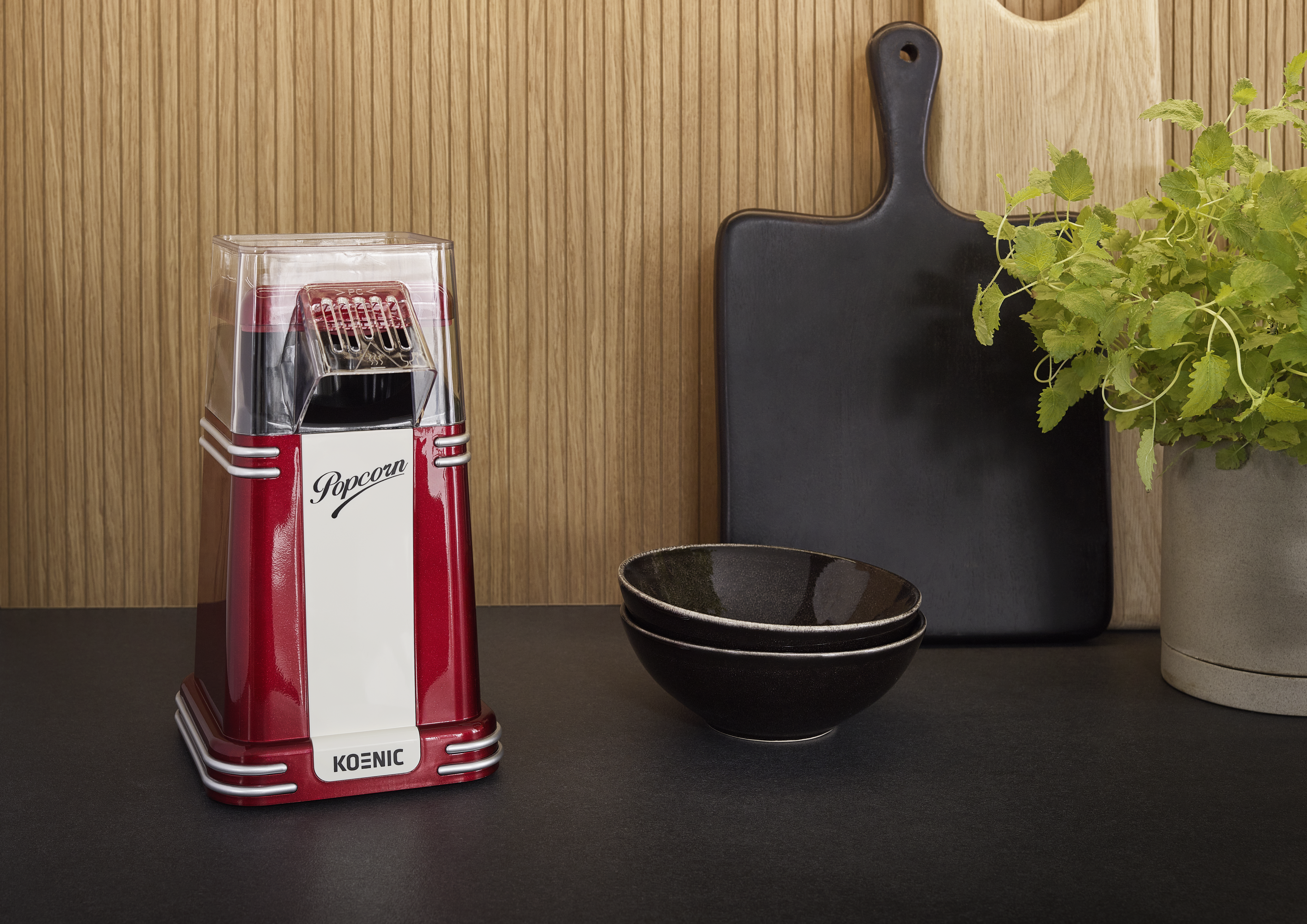 Roter und weißer Popcorn-Maker, schwarze Schalen, Schneidebretter und eine Topfpflanze stehen auf dunkler Oberfläche, Holz Hintergrund.