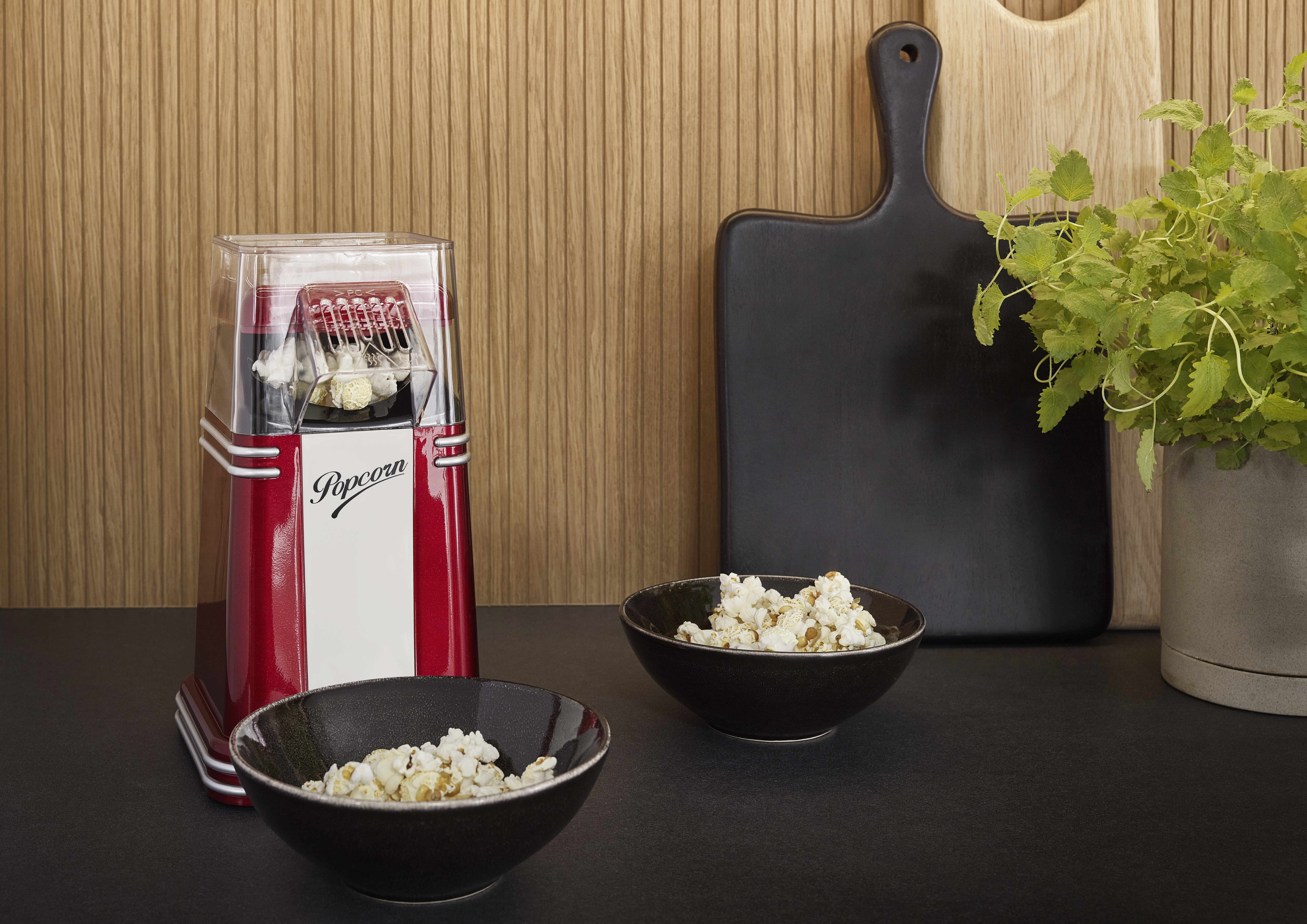 Ein rot-weißer Popcornbereiter, zwei Schalen Popcorn, Schneidebretter und eine Pflanze in einem Topf stehen auf einer schwarzen Oberfläche.