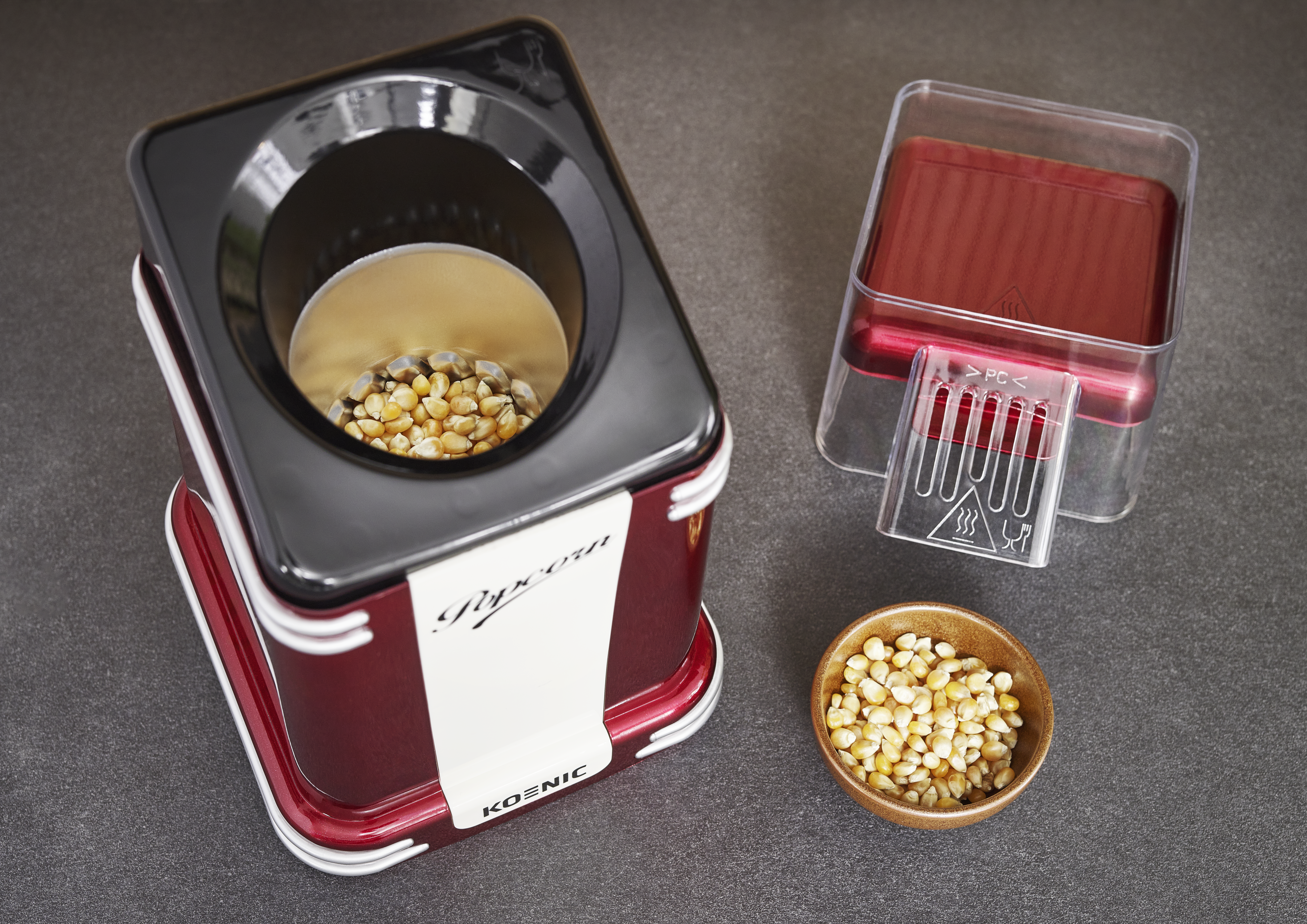 Ein rot-schwarzer Popcorn-Maker mit Maiskörnern im Inneren. Eine Schüssel mit Körnern und ein roter Deckel liegen daneben.