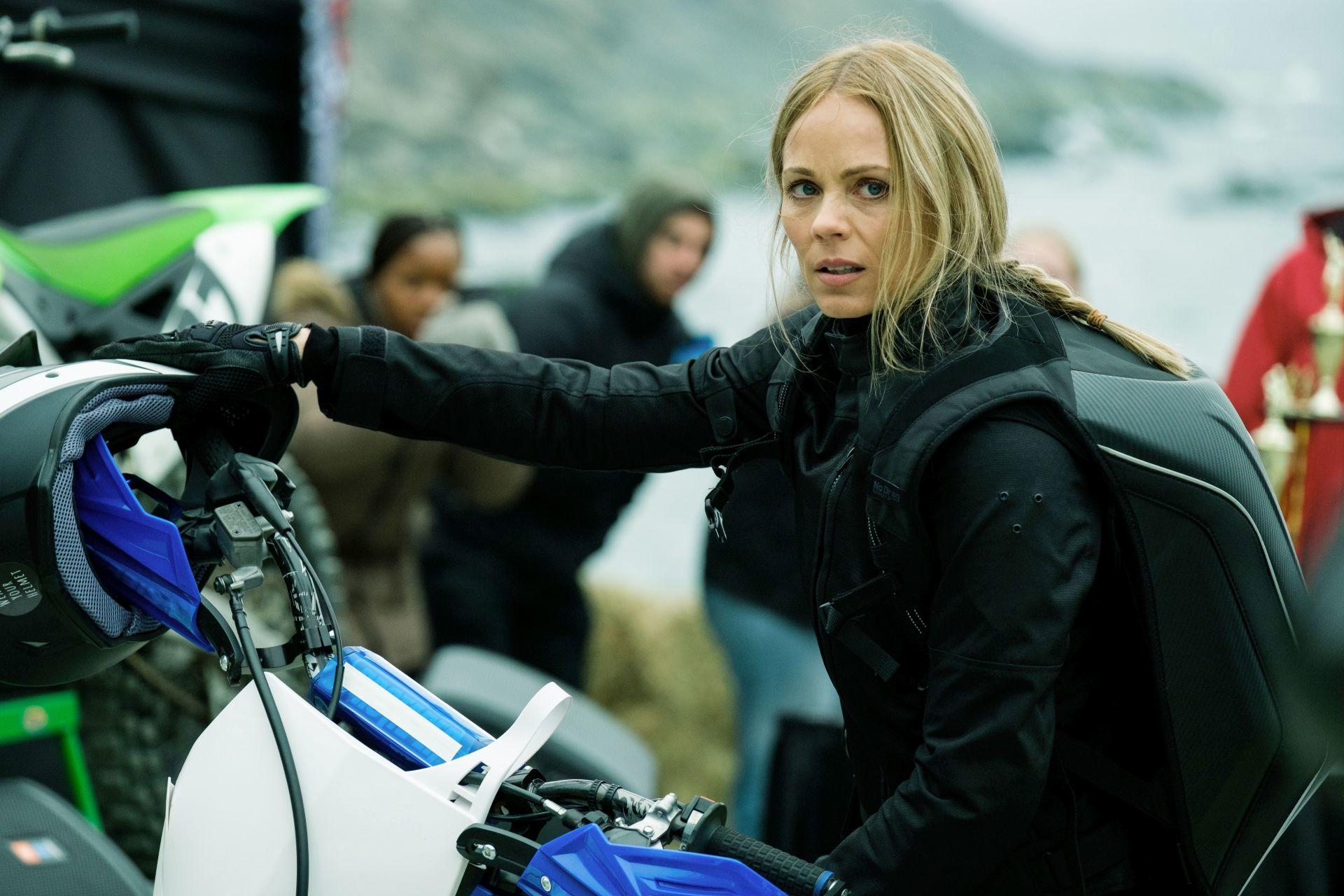 Frau in schwarzer Jacke an einem Motorrad. Sie hält den Helm mit besorgtem Blick. Andere sind im Hintergrund.