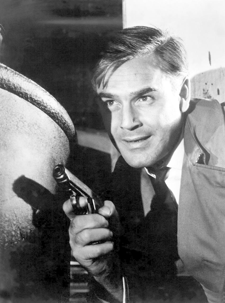 Ein Mann im Anzug hält eine Waffe und zielt auf ein unsichtbares Objekt. Er wirkt konzentriert und lächelt leicht. Schwarz-Weiß-Foto.