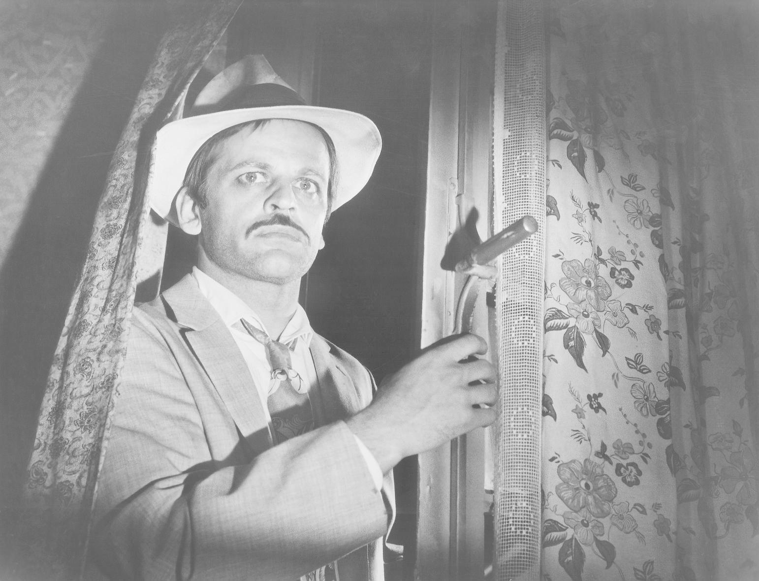 Ein Mann in Anzug und Hut hält ein Werkzeug an einem Fenster. Blumengardinen sind sichtbar. Die Szene scheint drinnen zu sein.