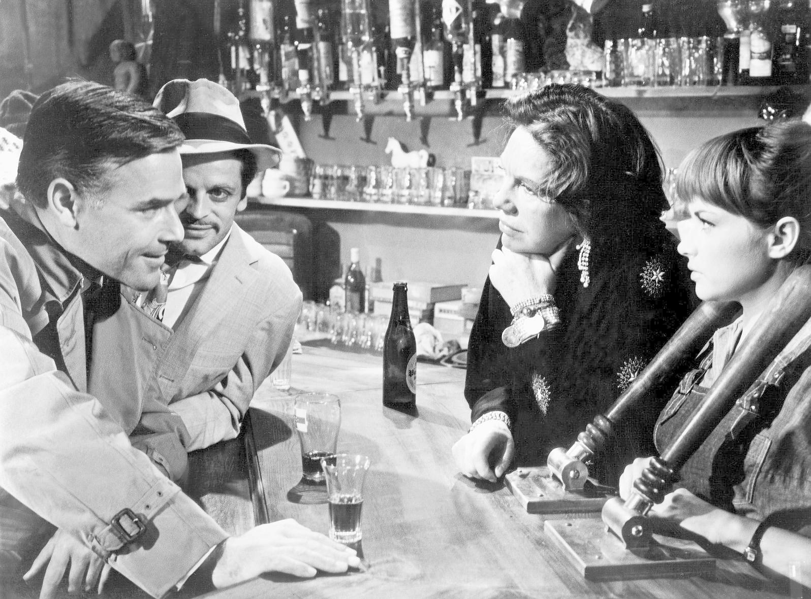 Vier Personen an einer Bar, einige schauen sich an, andere die Bar an. Flaschen und Gläser stehen auf der Bar. Schwarz-Weiß-Bild.