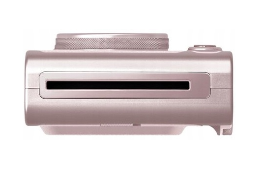 Pokazano różowo-złoty aparat natychmiastowy. Ma pokrętło obiektywu na górze, czarną szczelinę i białe tło.