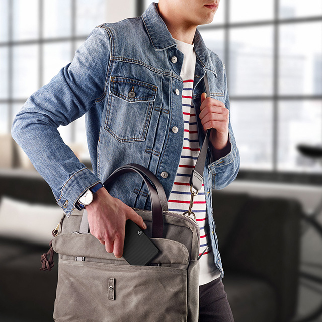 Un hombre con una chaqueta de mezclilla y una camisa a rayas mete un disco duro externo negro en una bolsa mensajera gris.