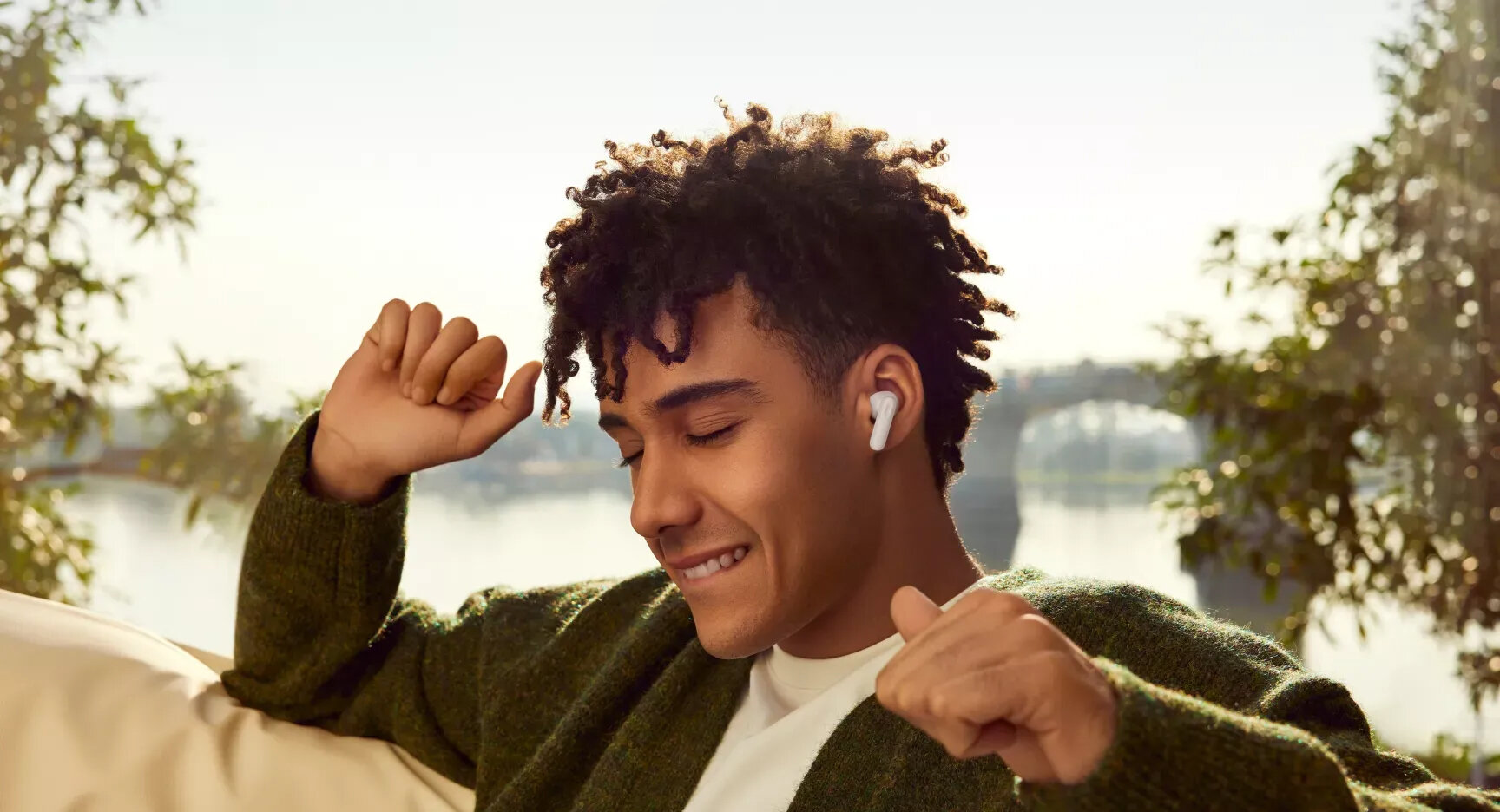 Mężczyzna uśmiecha się, nosząc słuchawki. Jest na zewnątrz, z mostem i wodą w tle, drzewami po obu stronach.