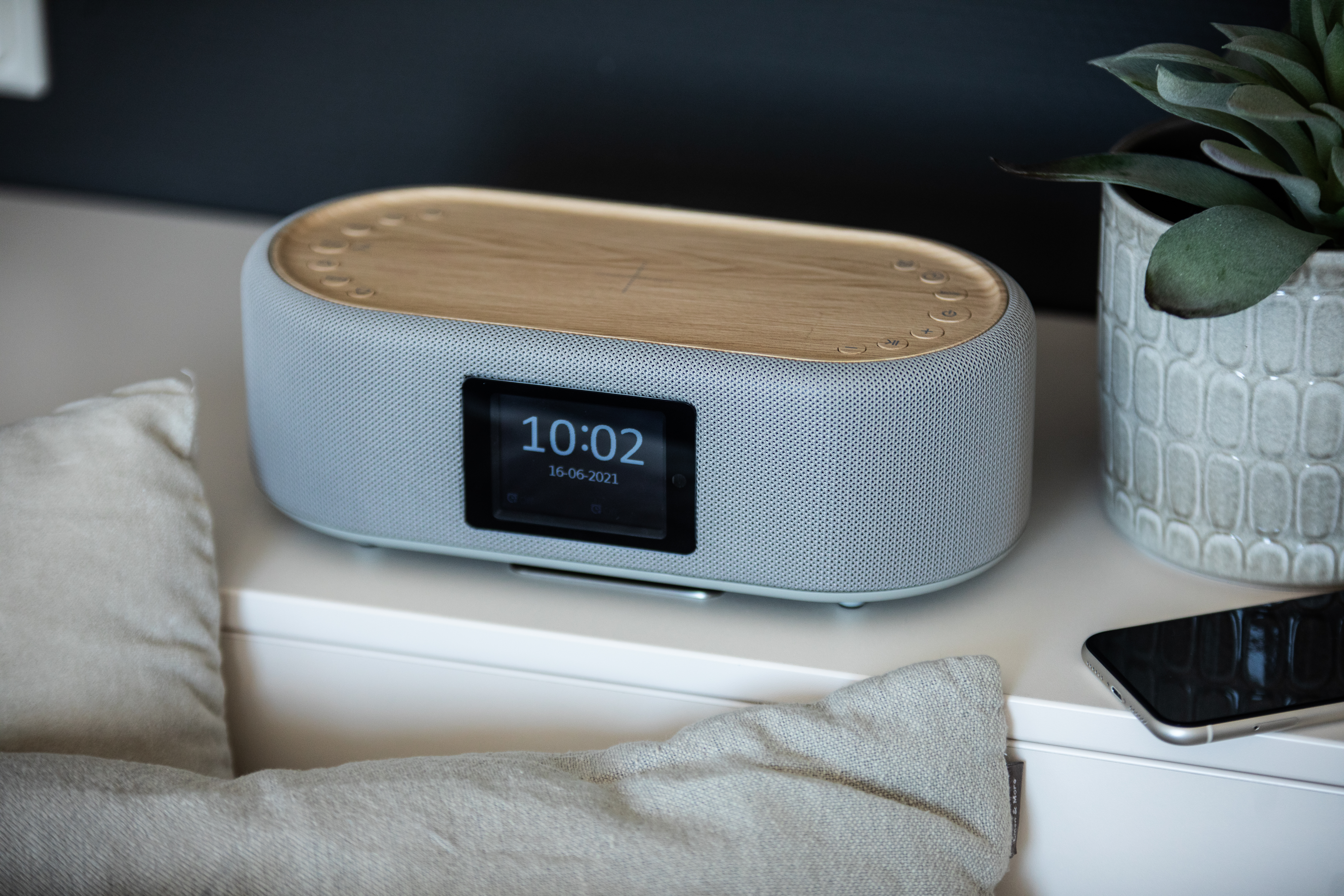 Szary głośnik z drewnianym blatem wyświetla godzinę. W pobliżu roślina w doniczce i telefon. Poniżej poduszka.