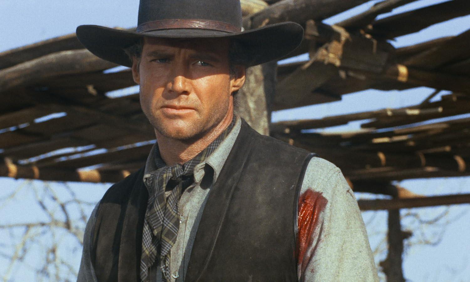 Ein Mann mit Cowboyhut und Weste, mit blutbeflecktem Hemd. Er starrt intensiv, im Hintergrund eine Holzkonstruktion.