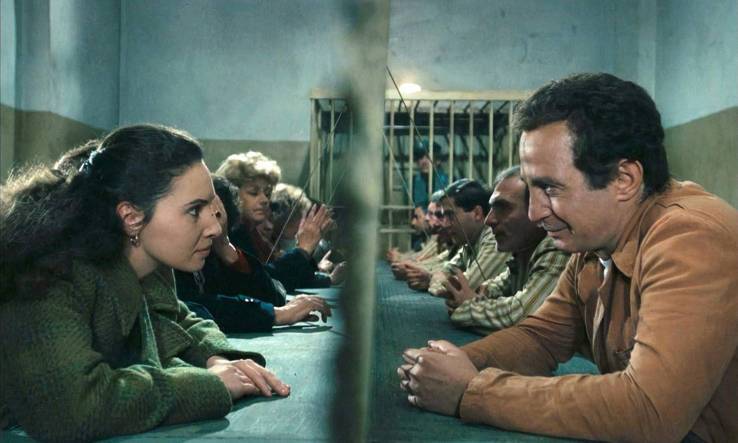 Menschen stehen sich an einem Tisch gegenüber, getrennt durch Glas. Auf der einen Seite eine Frau in Grün, auf der anderen Männer im Gefängnis.