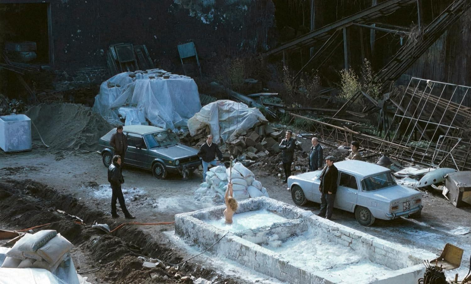 Eine Frau in einem Becken mit milchigem Wasser, umgeben von Menschen und Autos. Die Szene hat eine industrielle, etwas heruntergekommene Umgebung.