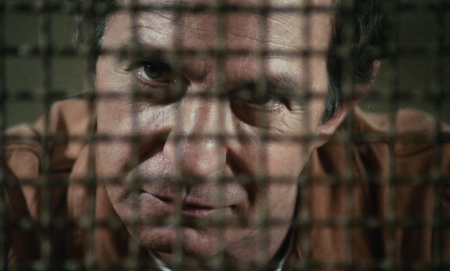 Das Gesicht eines Mannes, durch ein Metallgitter gesehen. Er trägt ein orangefarbenes Hemd. Der Mann hat graues Haar und einen ernsten Gesichtsausdruck.