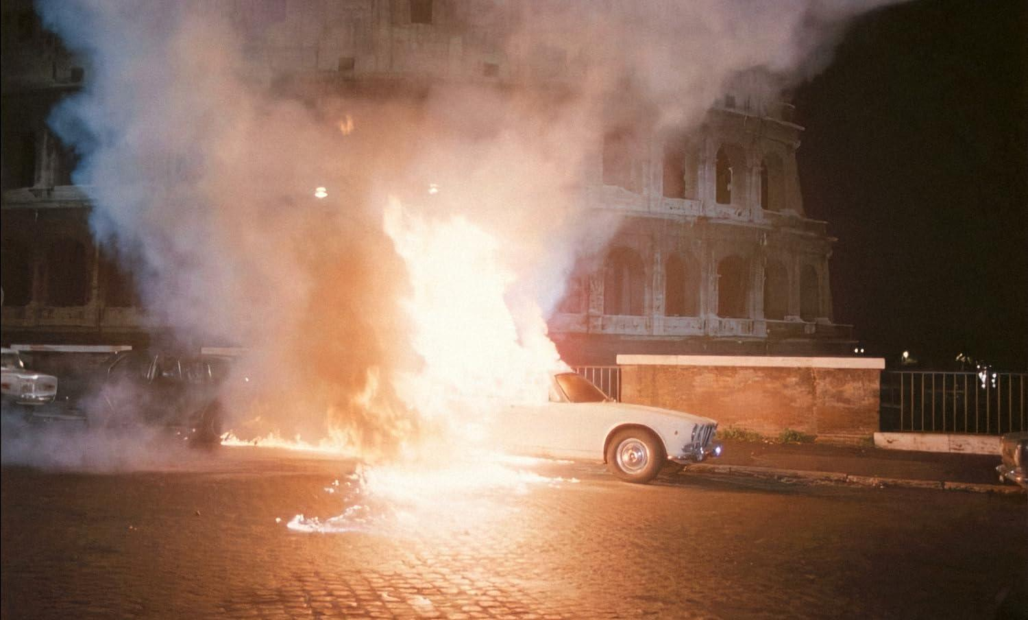Ein weißes Auto brennt vor einem Gebäude bei Nacht. Flammen und Rauch steigen auf. Im Hintergrund ist das Kolosseum zu sehen.