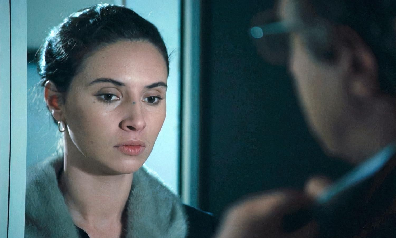 Eine Frau mit dunklem Haar blickt nachdenklich. Ein Mann ist rechts unscharf. Die Frau trägt einen grauen Mantel.