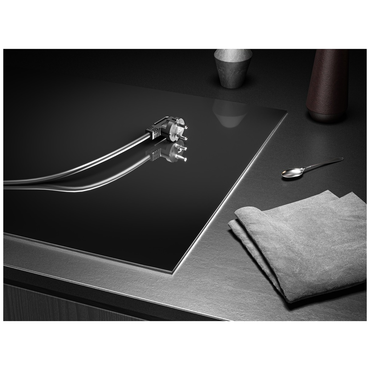 A black hob with an electrical plug, spoon, grey cloth, and decorative items on a dark surface. Good lighting.