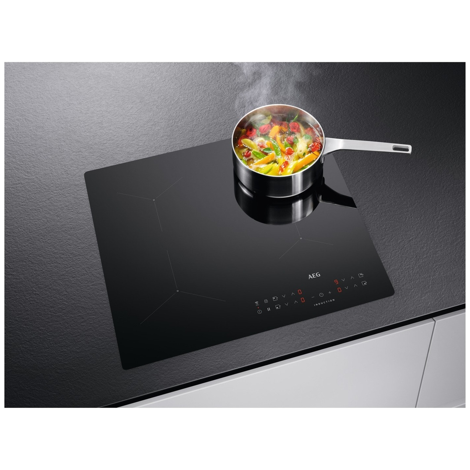 A black induction cooktop with a pot of vegetables steaming on it. The cooktop has a digital display and the AEG logo.