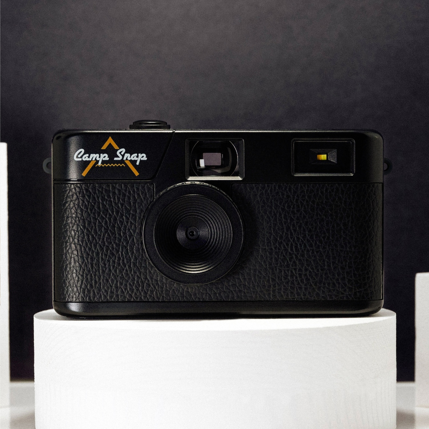 A black camera labeled 'Camp Snap' is on a white podium against a dark background. The camera has a textured leather grip.