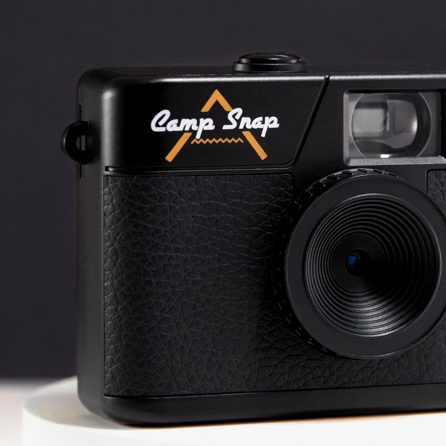 A black Camp Snap camera is on a white surface with a dark background. The camera has a lens, viewfinder and the brand logo.