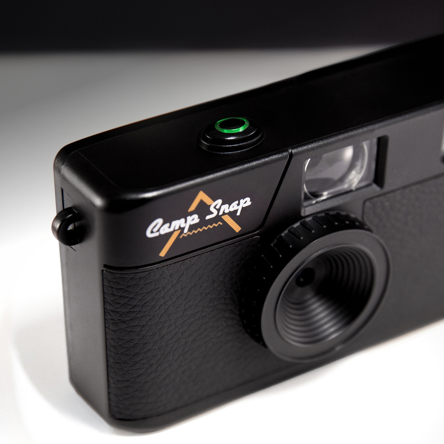 A black Camp Snap camera is on a white surface. It has a green button and a lens. The logo is gold and white.