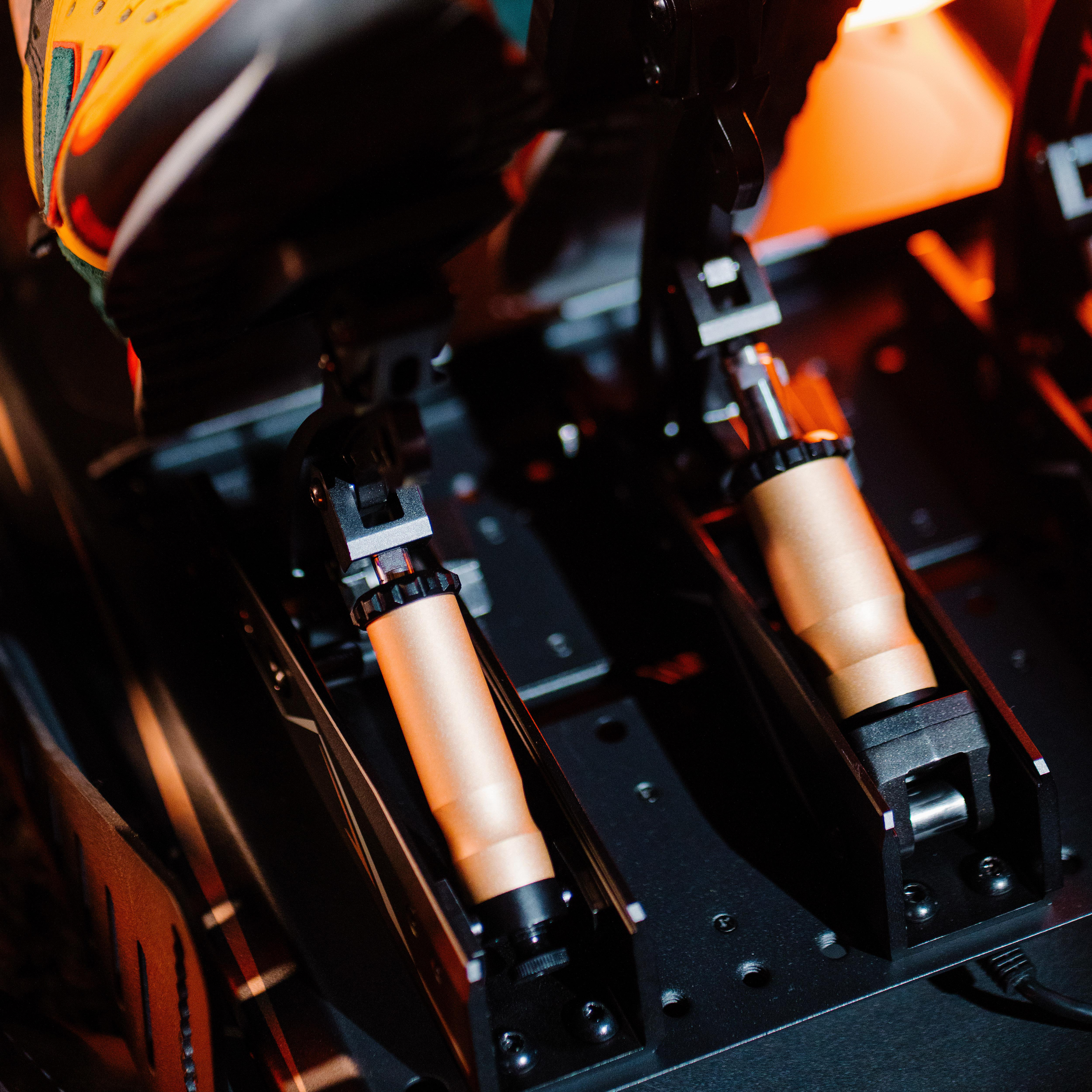 Nahaufnahme von Rennpedalen. Goldzylinder mit schwarzen Details sind auf einer schwarzen Platte montiert. Ein oranger Schuh ist sichtbar.
