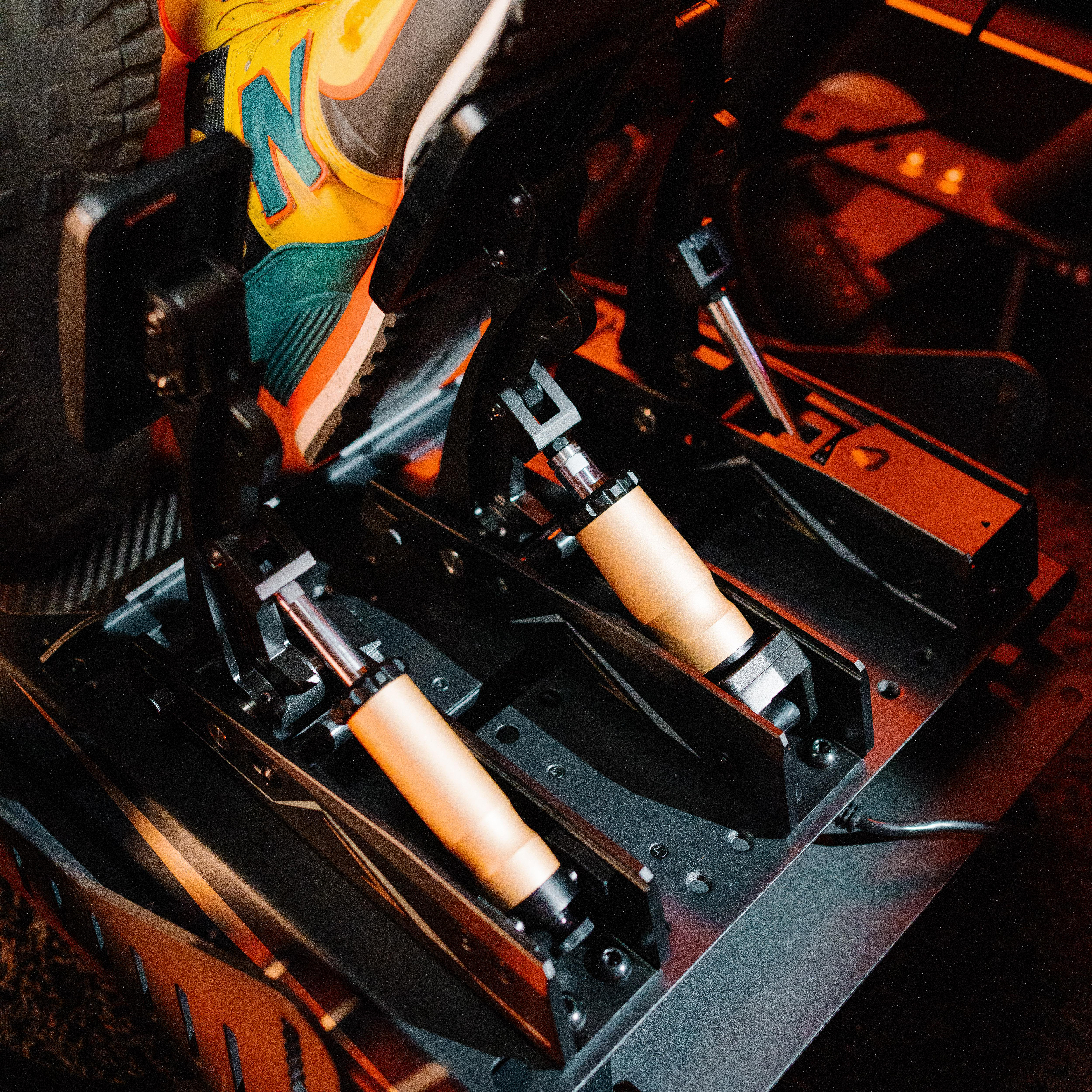 Nahaufnahme von Rennsimulatorpedalen mit orangefarbenen und schwarzen Details. Ein Fuß mit buntem Schuh ist teilweise sichtbar.