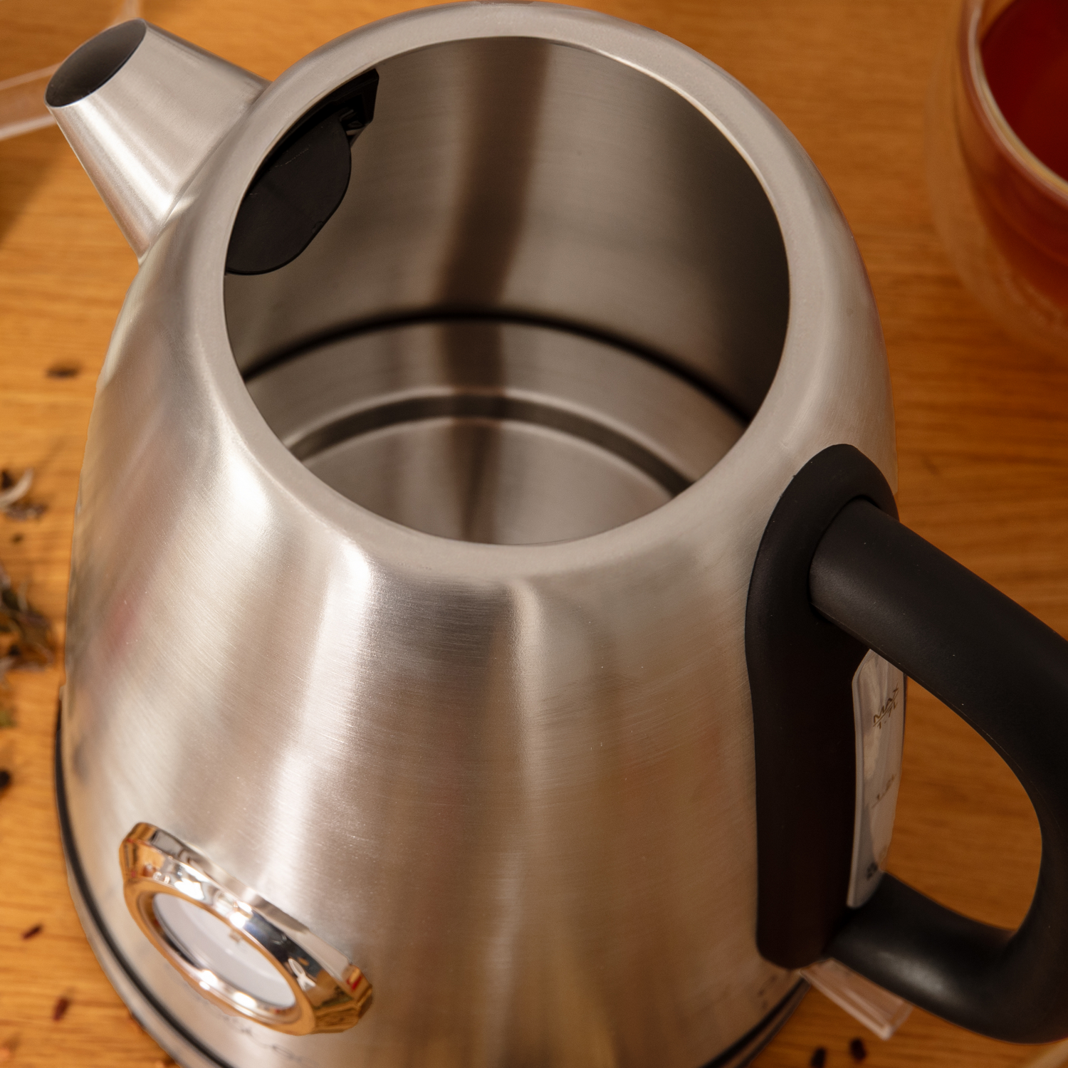 Una tetera eléctrica de acero inoxidable con asa negra, sobre una superficie de madera. En el fondo se ve una taza de té.