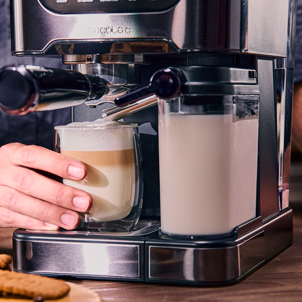 Máquina de espresso dispensando leche en un vaso de doble pared. Una mano sostiene el vaso. Contenedor de leche a la derecha.