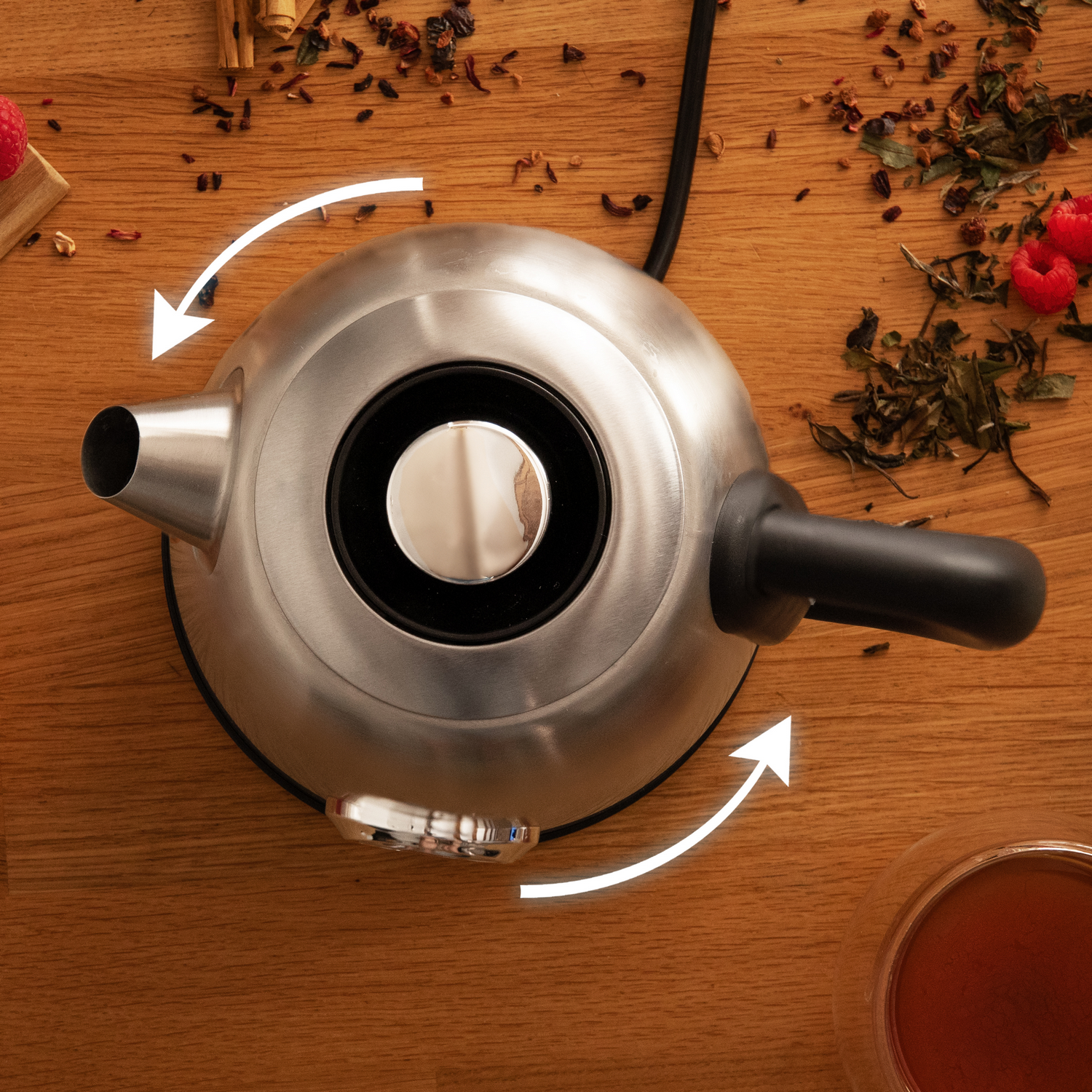 Vista aérea de una tetera plateada sobre una mesa de madera con hojas de té, frambuesas y un vaso de té.