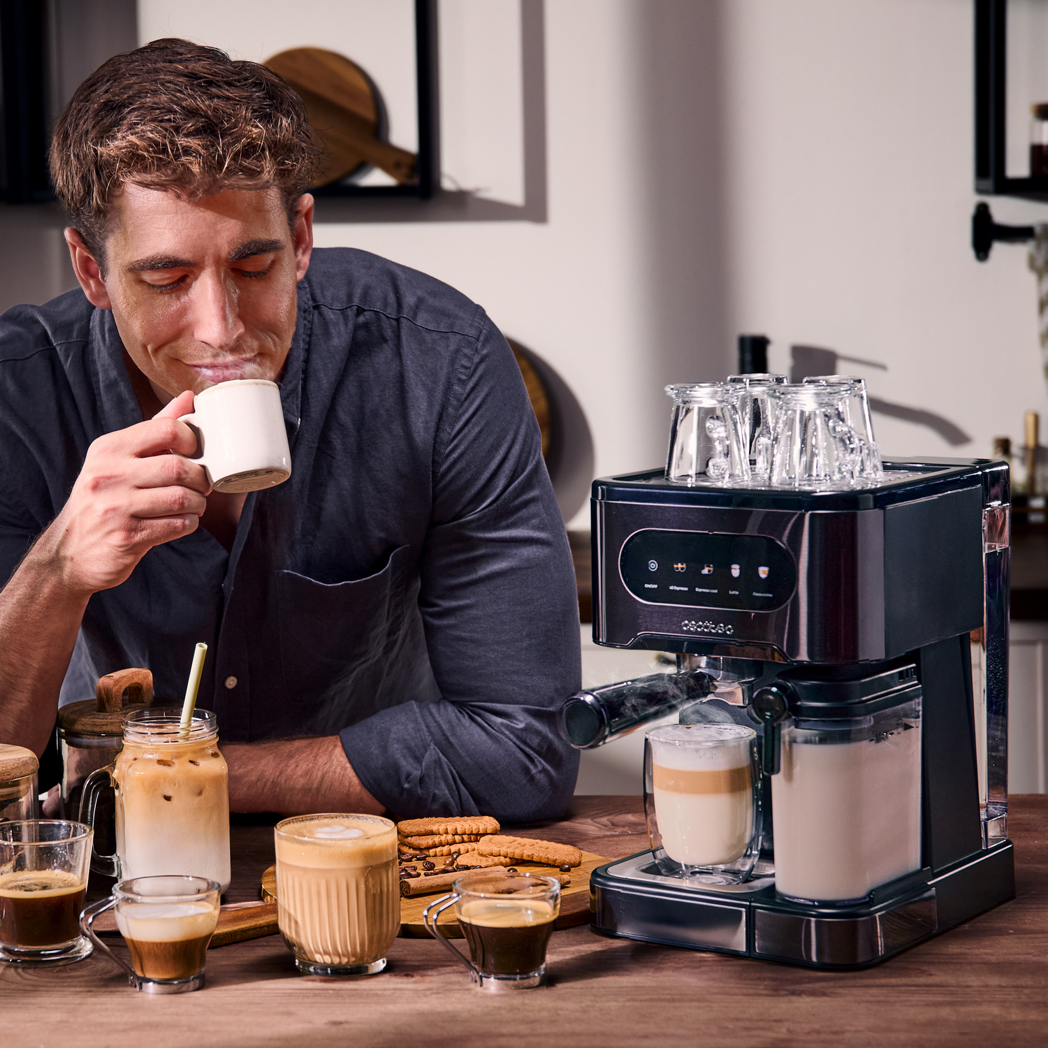 Hombre oliendo café de una taza. Cafetera, varias bebidas de café y galletas en una mesa de madera.