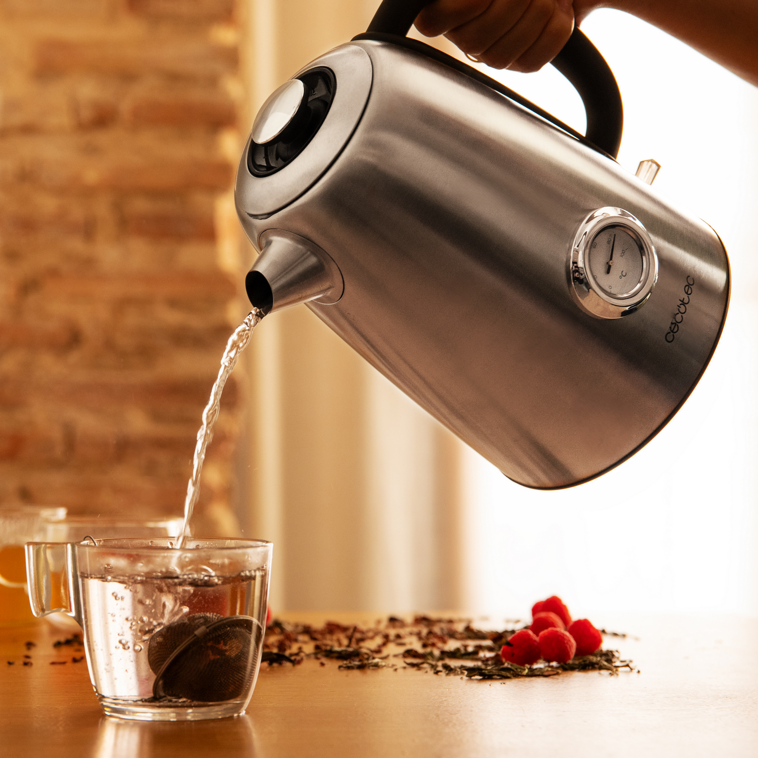 Una tetera de acero inoxidable vierte agua en una taza de vidrio con un infusor de té. Las frambuesas y las hojas de té están esparcidas.