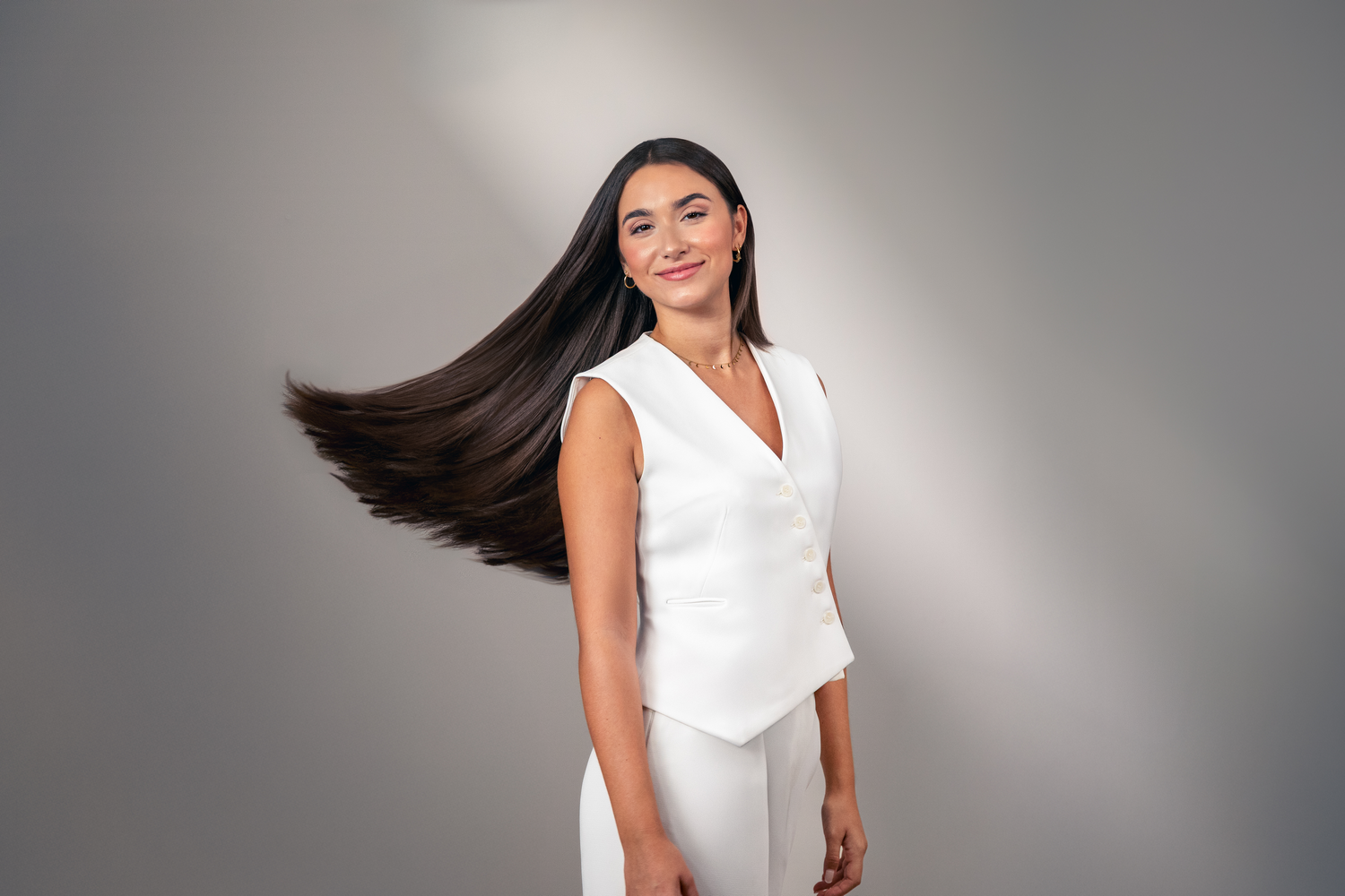 Una mujer con cabello largo y oscuro, vestida con chaleco y pantalón blancos, sonríe. Su pelo fluye con el viento.