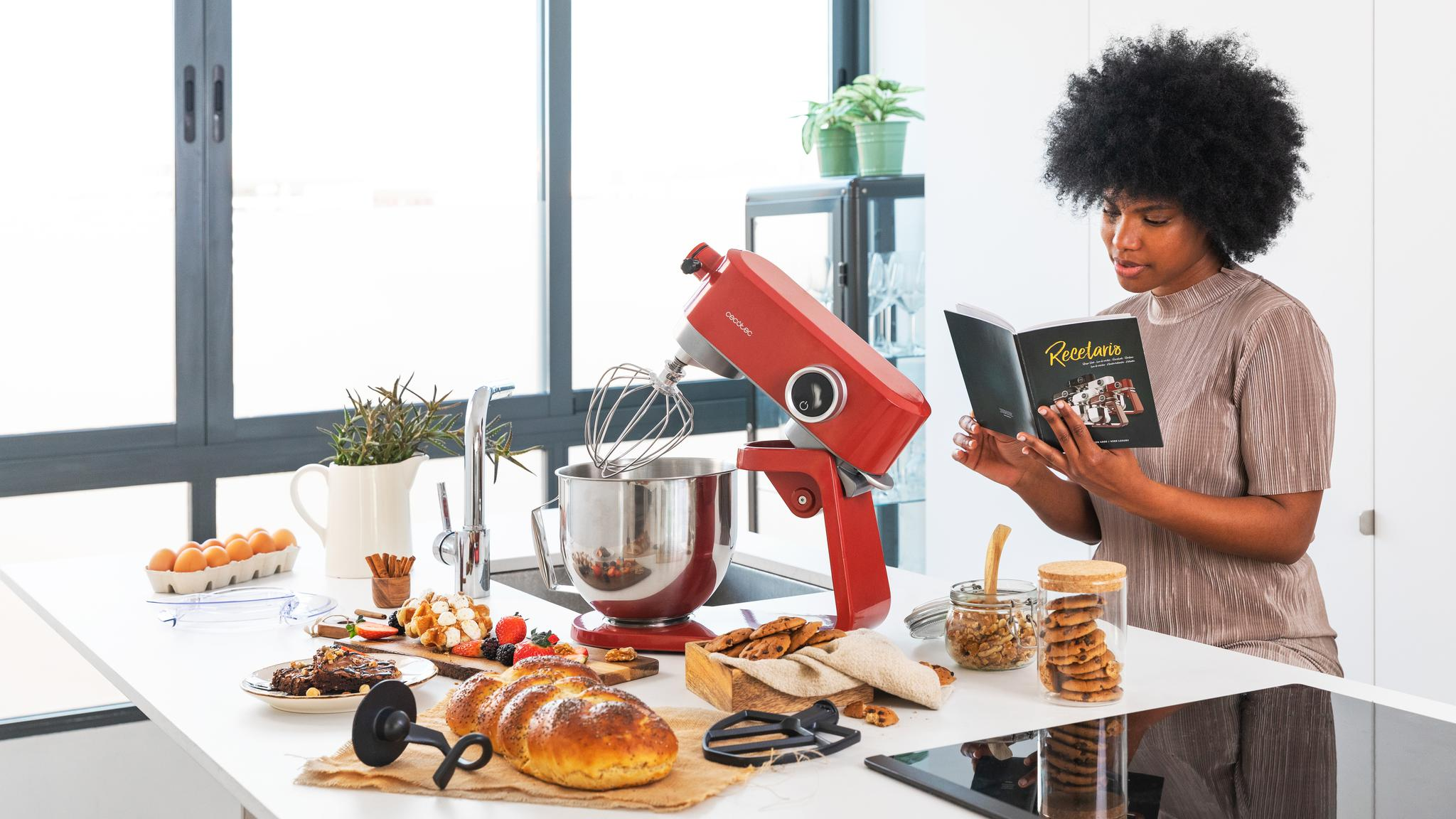 Mujer lee un libro de cocina en una cocina con una batidora roja y varios productos horneados en la mesa.