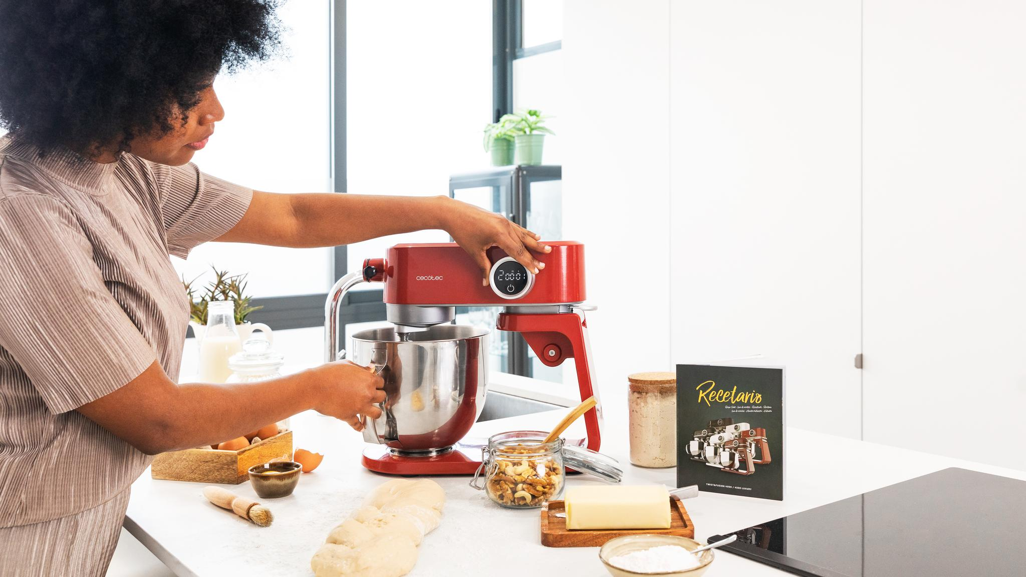Una mujer usa una batidora de cocina roja. Varios ingredientes para hornear están en la encimera: huevos, harina, mantequilla, nueces y un libro de recetas.