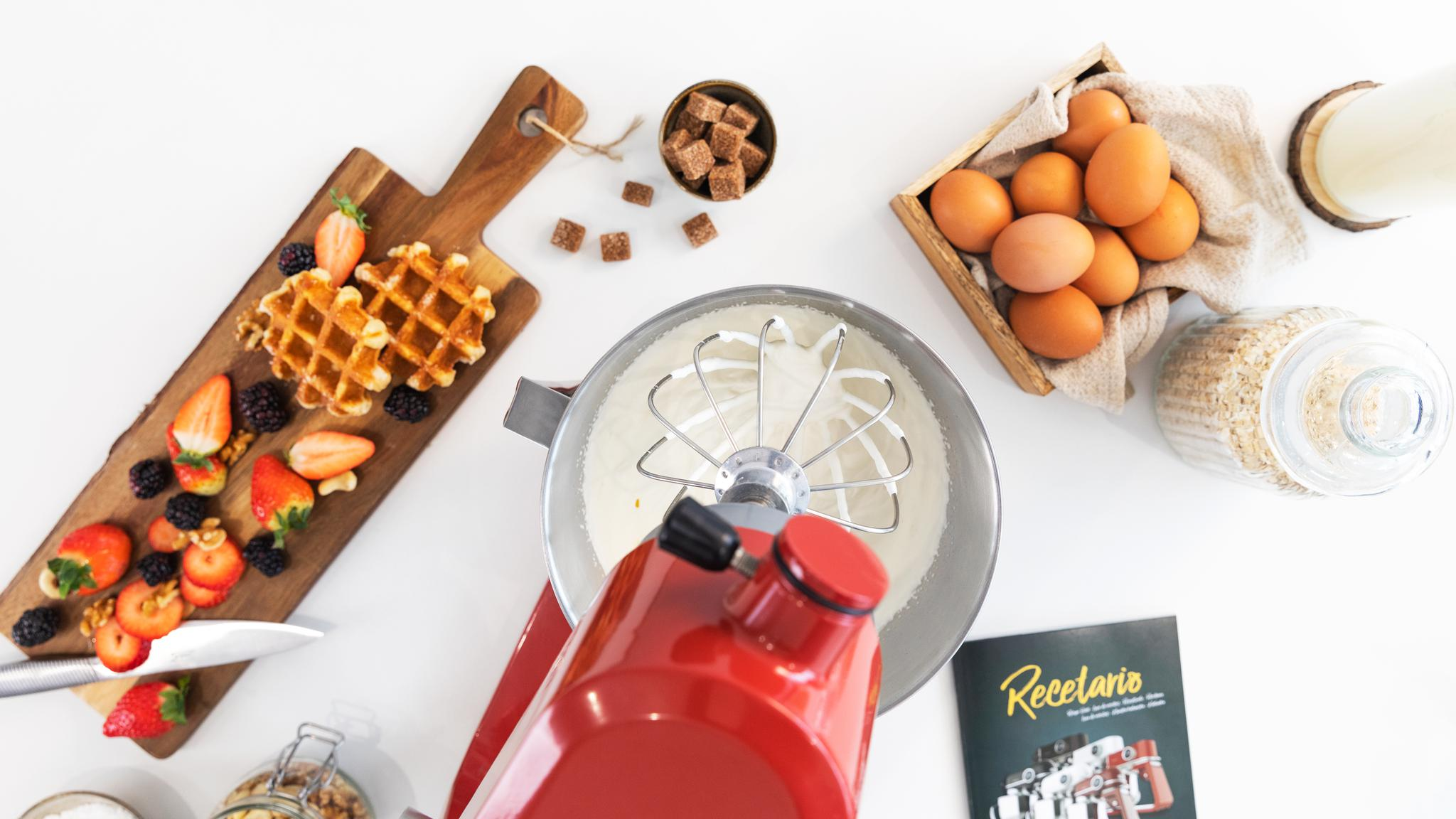 Toma cenital de una escena de cocina con gofres, bayas, una batidora, huevos y azúcar. Ingredientes de horneado.