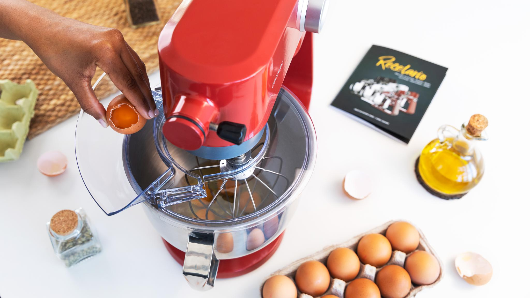 Una persona rompe un huevo en una batidora roja. Otros huevos, aceite y un libro de cocina están sobre una superficie blanca.