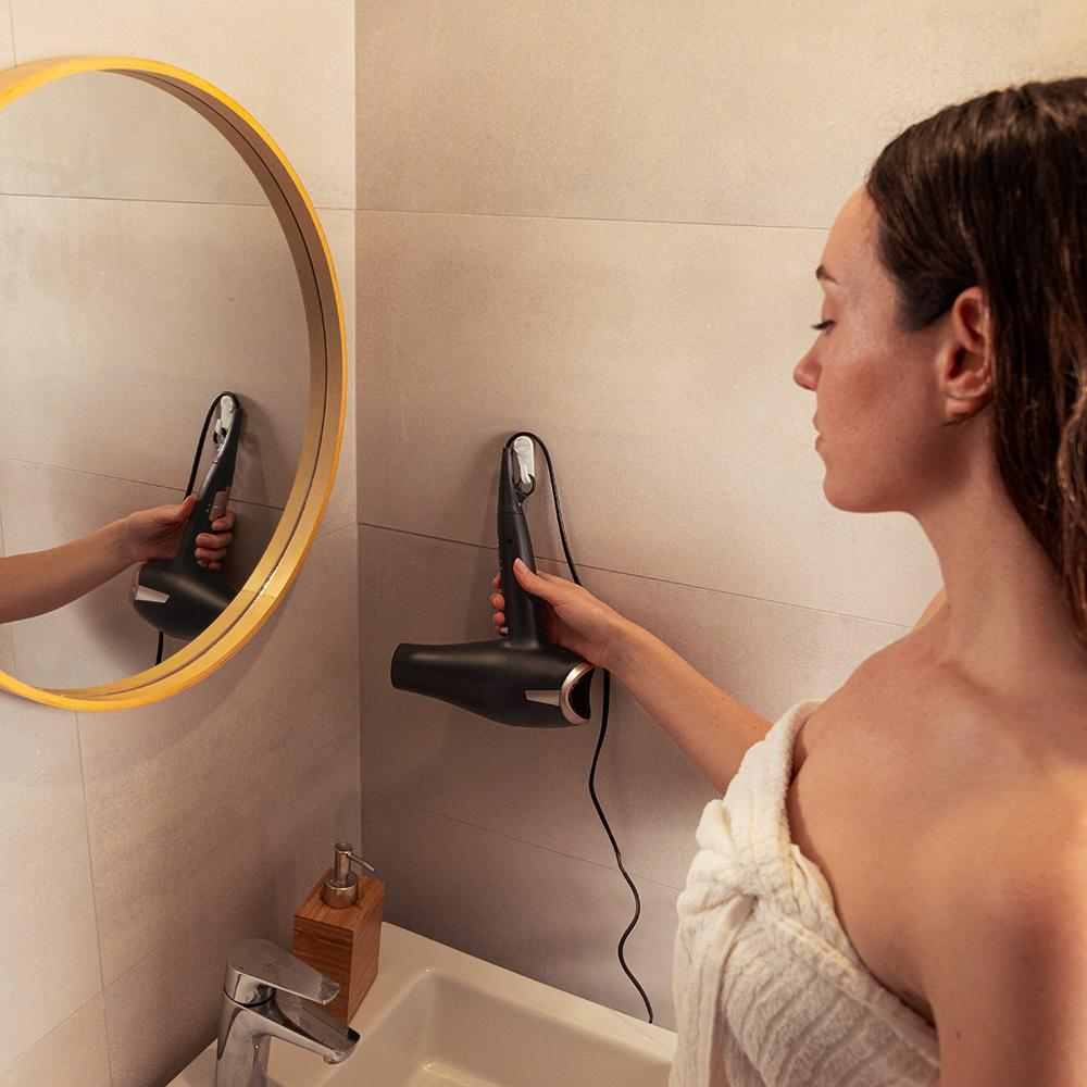 Mujer en el baño con secador de pelo. El espejo refleja el secador. Toalla blanca, lavabo y dispensador de jabón también visibles.