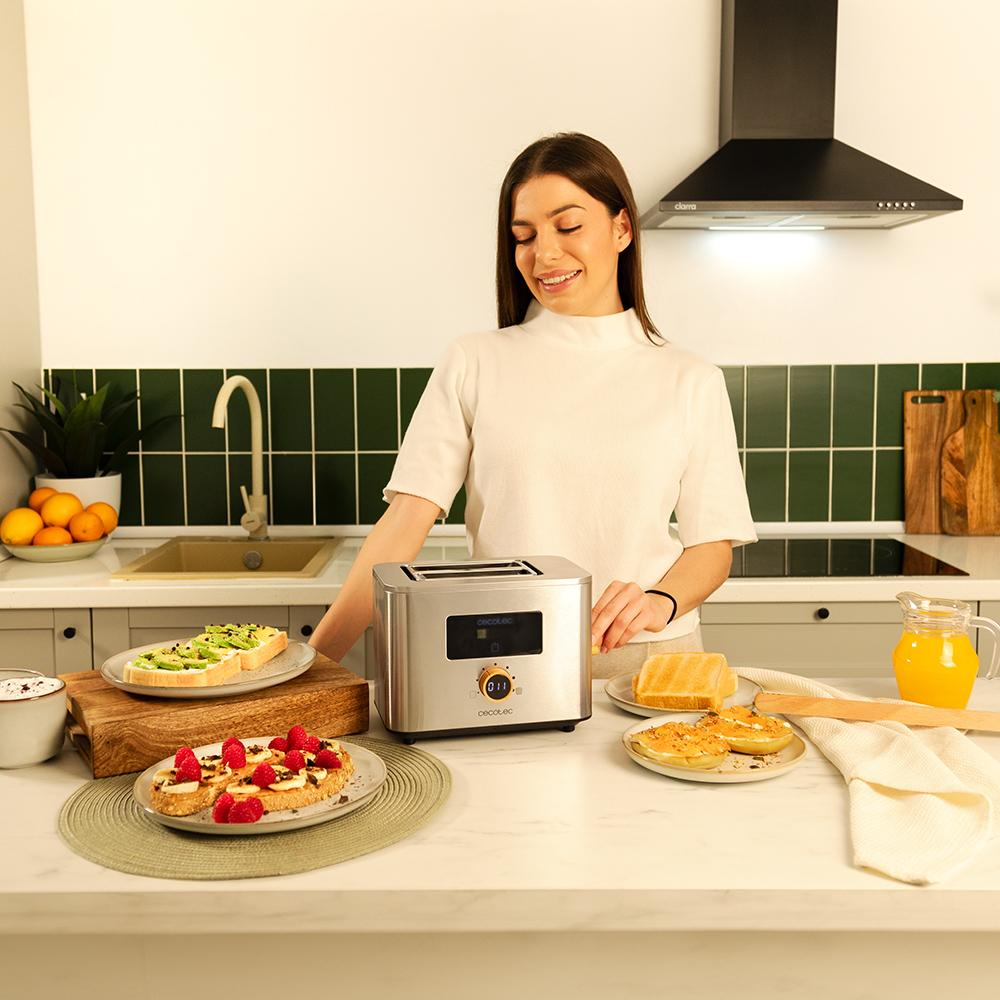 Una mujer sonríe en una cocina, cerca de una tostadora, tostadas y platos de comida. Pared de azulejos verdes en el fondo.