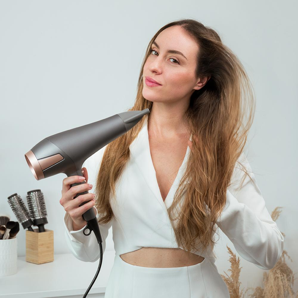 Mujer de blanco se seca el pelo con un secador gris. Cepillos de pelo y planta en el fondo. Habitación luminosa.