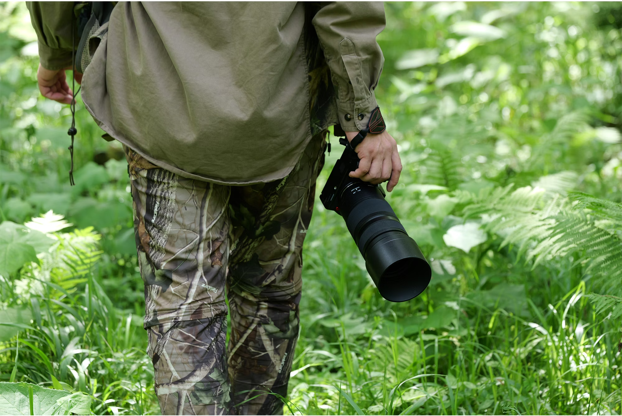 Une personne en pantalon camouflage et chemise beige tient un gros appareil photo dans une forêt avec un feuillage vert.