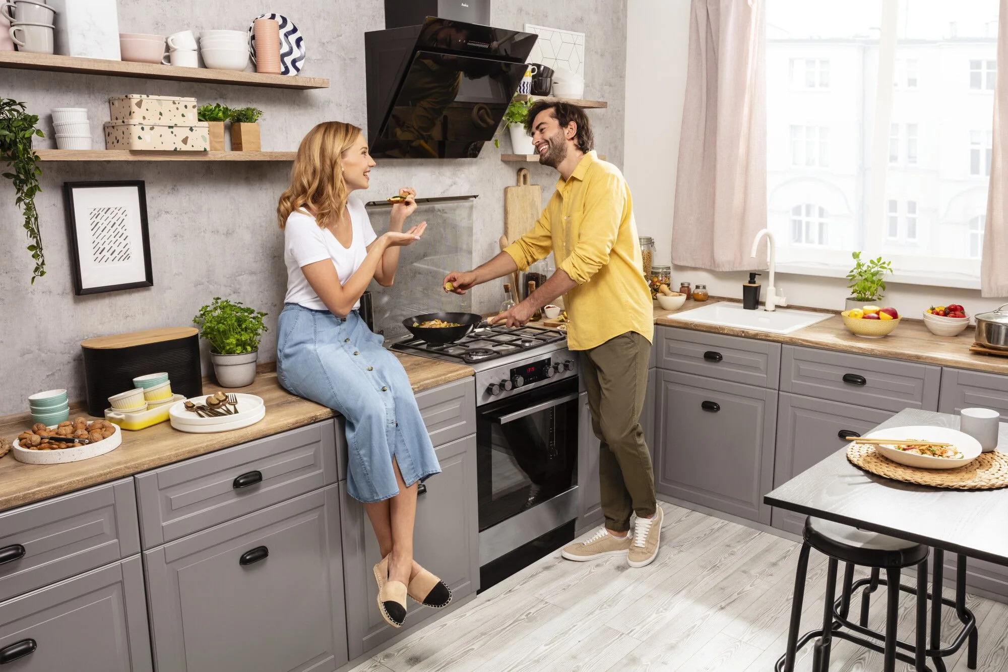 Para w nowoczesnej kuchni. Kobieta siedzi na blacie, mężczyzna gotuje na kuchence. Światło słoneczne wpada przez okno.