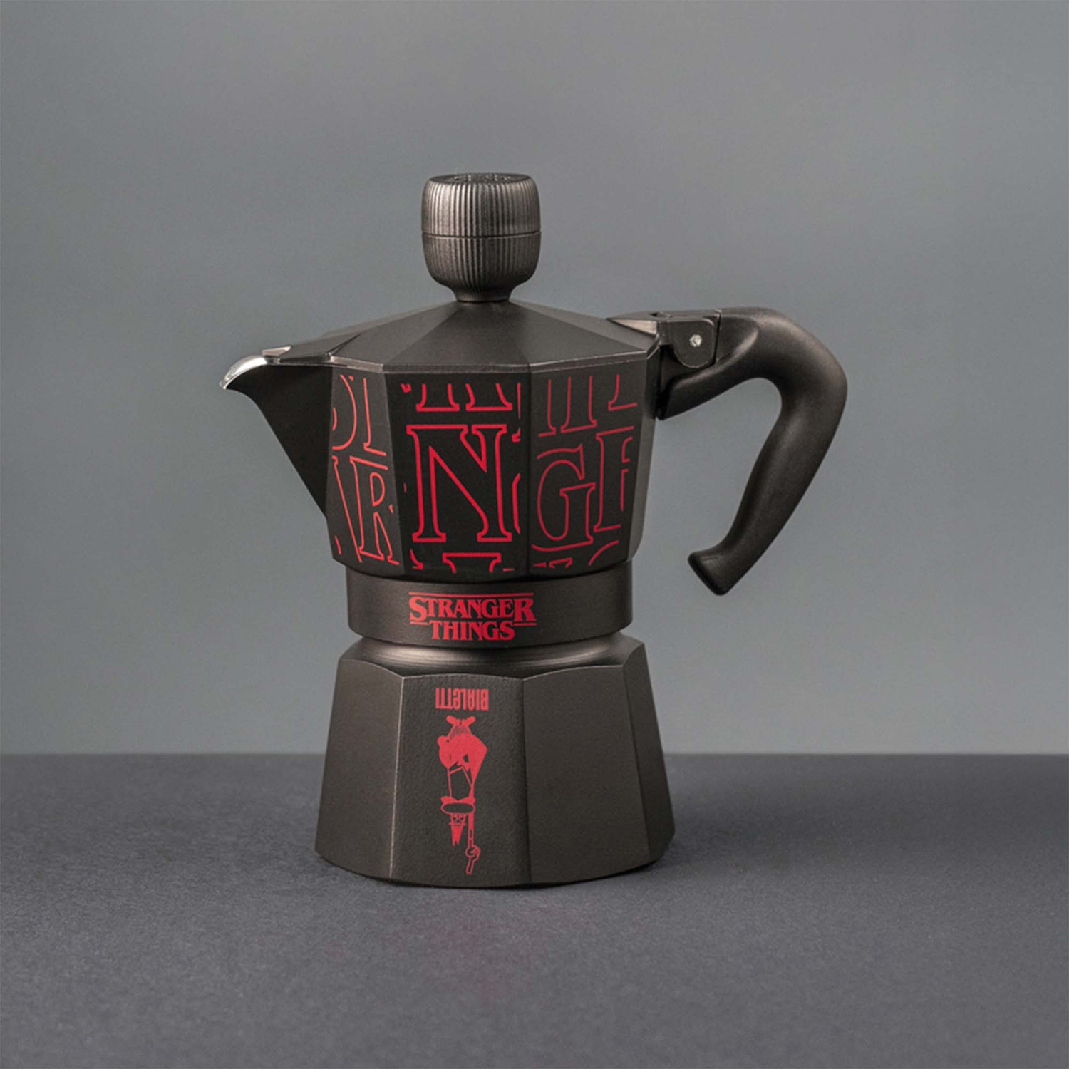 Caffettiera nera con logo rosso 'Stranger Things'. È su una superficie scura e su sfondo grigio.
